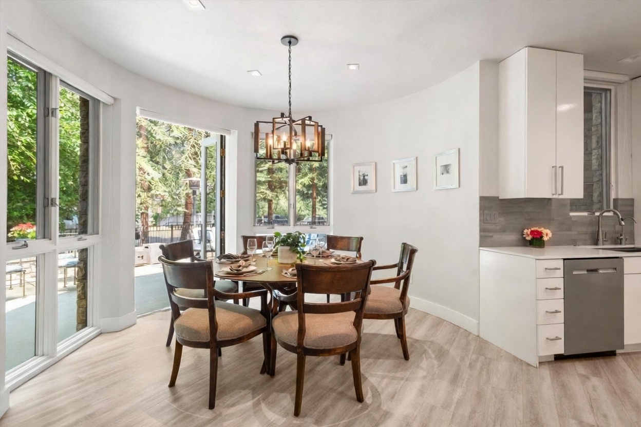 This bright dining area features a round wooden table surrounded by six chairs with upholstered seats, positioned under a modern geometric chandelier. The space is open to a kitchen area with white cabinetry and a stainless steel dishwasher, and it includes large windows and glass doors that provide a view of the outdoor greenery. The light-toned wood-look flooring and neutral walls create a clean, inviting atmosphere suitable for casual or formal dining.