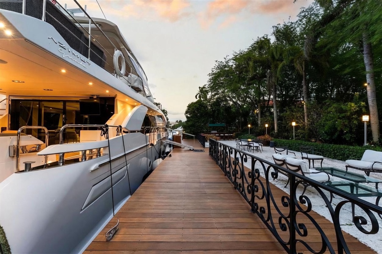 This image captures a luxurious waterfront scene featuring a large yacht docked alongside a wooden pier. To the right of the pier, a landscaped patio area is furnished with outdoor seating, set against a backdrop of lush tropical trees and warm evening lighting. The perspective is low and wide, emphasizing the seamless transition between the private dock and the elegant outdoor living space.