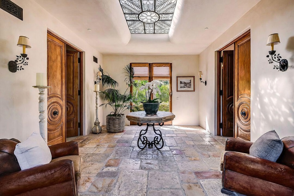 This elegant foyer features a rustic stone floor and a dramatic skylight with intricate metal detailing that casts patterned shadows across the space. Two matching leather armchairs flank the entrance, while a central stone-topped table holds a potted orchid, creating a sophisticated and welcoming atmosphere. The room is framed by symmetrical wooden doors and wall-mounted sconces, offering a balanced and refined architectural aesthetic.