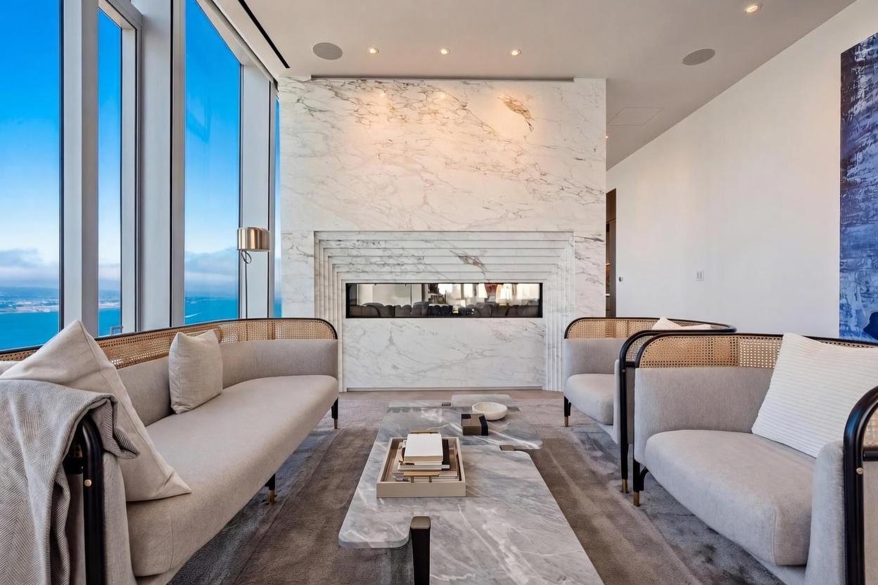 This sophisticated living room features a striking stone-clad chimney breast that serves as a central focal point, flanked by expansive floor-to-ceiling windows offering panoramic water views. The seating area is arranged with two curved, cane-backed sofas and a tiered stone coffee table, all set upon a plush neutral rug. The space exudes a modern, high-end aesthetic with a clean, minimalist color palette and refined textures.