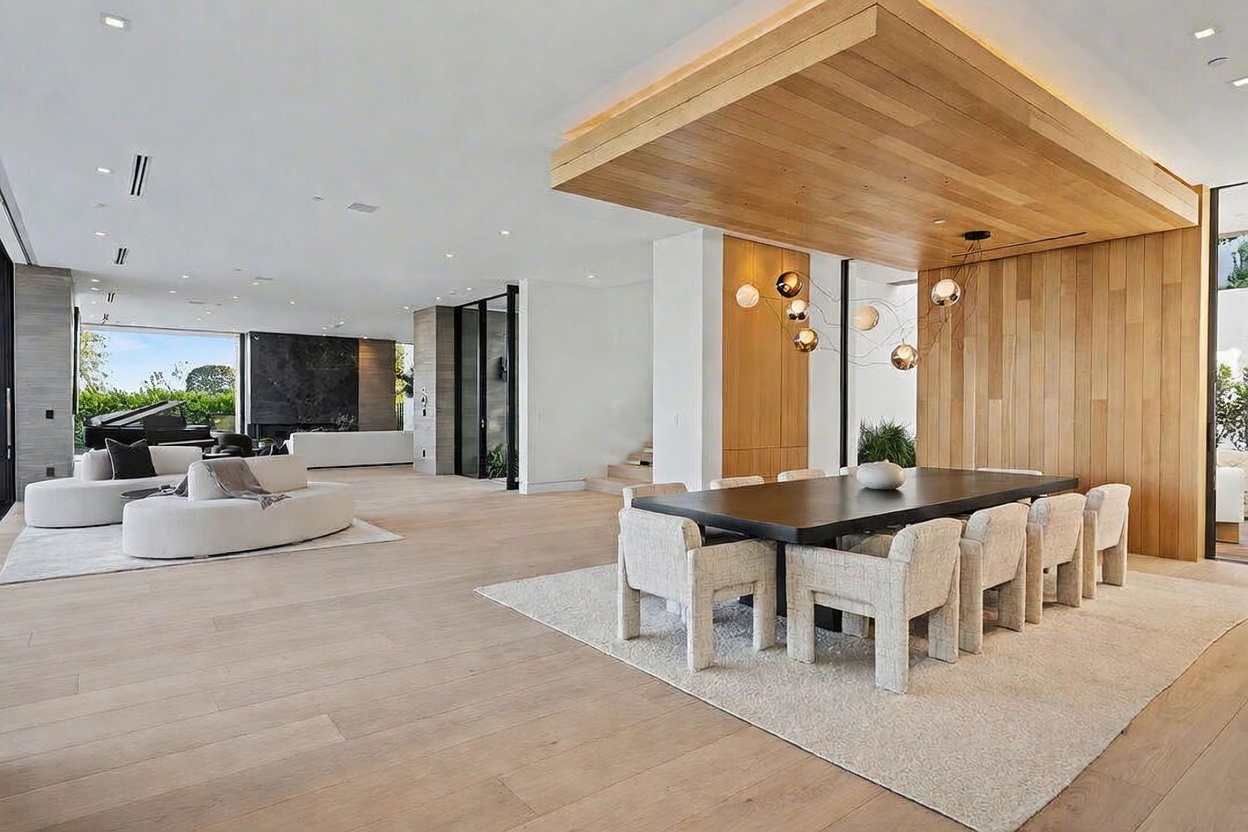 This expansive open-concept living and dining area showcases a sophisticated modern aesthetic with light wood flooring and a neutral color palette. The dining space is defined by a dark rectangular table surrounded by eight textured chairs, positioned beneath a striking wooden ceiling feature with integrated lighting. In the background, a curved white sofa sits in the living area, which features a dark stone wall accent and large windows that provide a seamless connection to the outdoor environment.