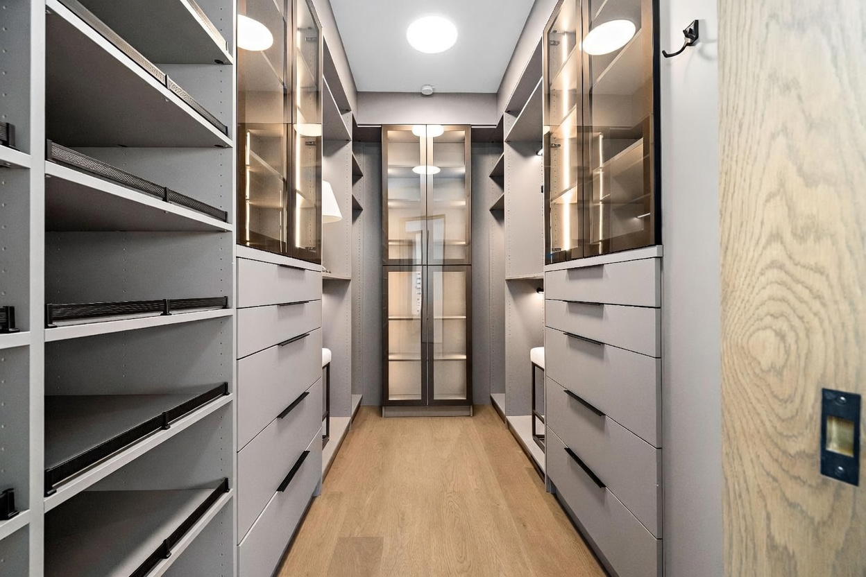 This sophisticated walk-in closet features a symmetrical layout with light gray cabinetry, integrated lighting, and glass-fronted display cabinets. The space is anchored by light wood flooring and a central glass-enclosed storage unit, creating a clean, organized, and high-end aesthetic. The perspective is a straight-on shot looking down the center aisle, emphasizing the depth and organized storage capacity of the room.