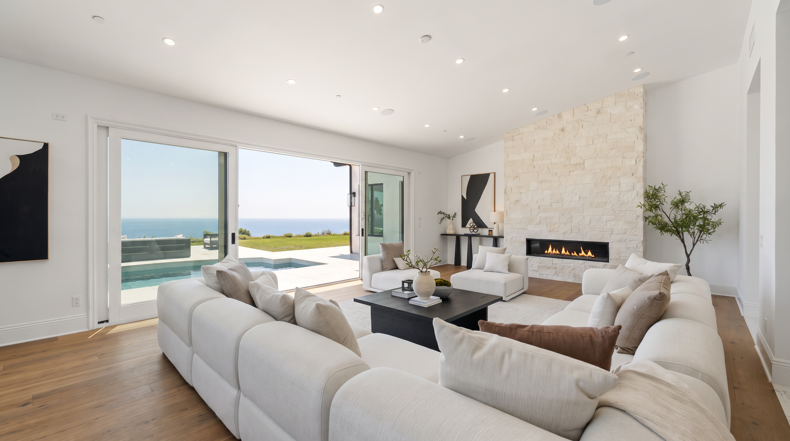 This bright and airy living room features a large, light-colored sectional sofa arranged around a dark coffee table, creating a comfortable seating area. A prominent stone-clad chimney breast serves as a focal point, housing a linear fireplace, while expansive sliding glass doors open to reveal a patio, swimming pool, and ocean views. The space is finished with light wood flooring and recessed lighting, offering a seamless transition between indoor and outdoor living.