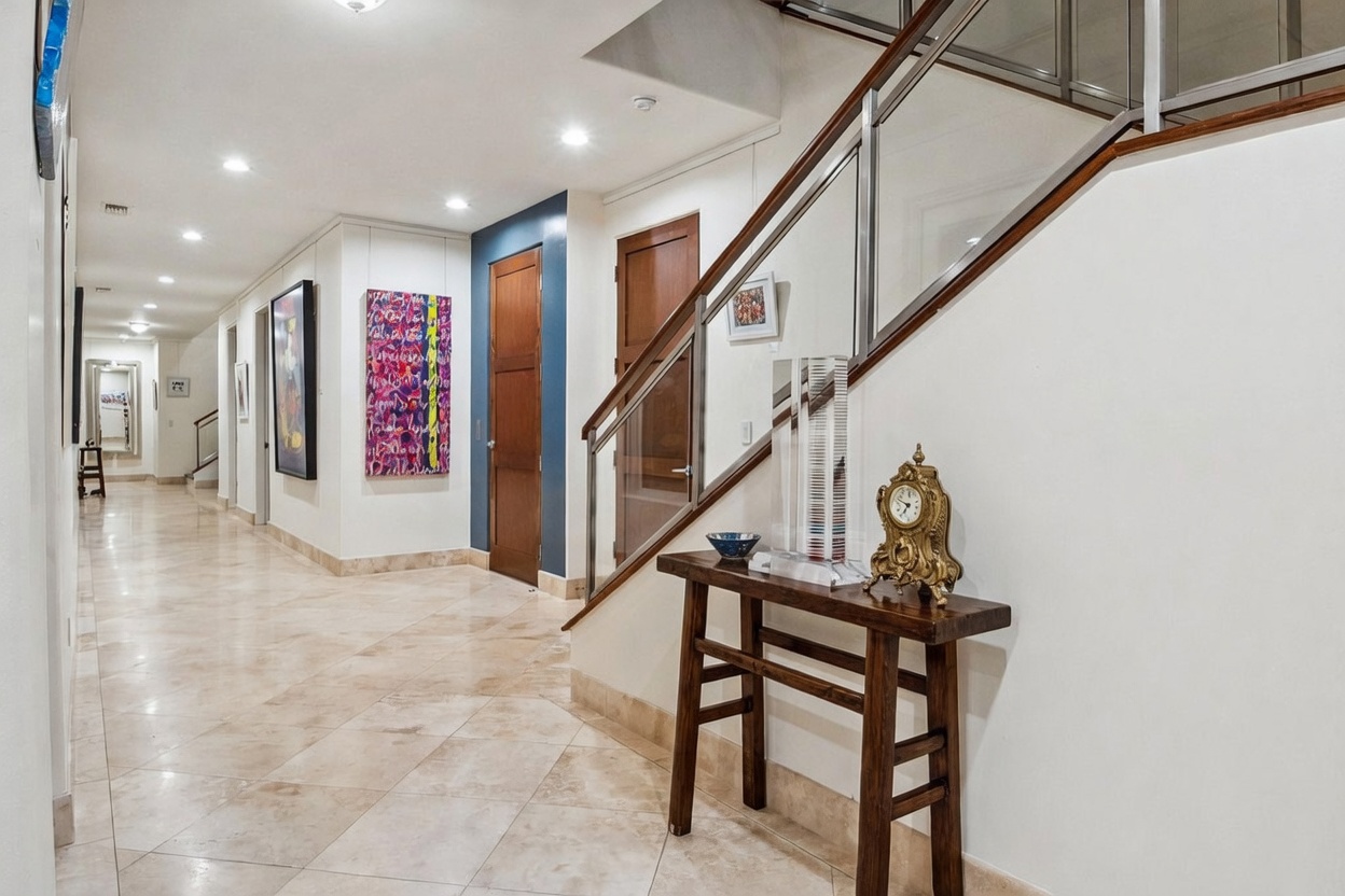 This interior view captures a bright, expansive hallway featuring light-colored stone flooring and a staircase with a modern glass railing. The space is decorated with vibrant abstract artwork and a wooden console table topped with a decorative clock, creating a sophisticated gallery-like atmosphere. The perspective looks down the long corridor, highlighting the clean lines and open transition between the lower level and the upper floor.