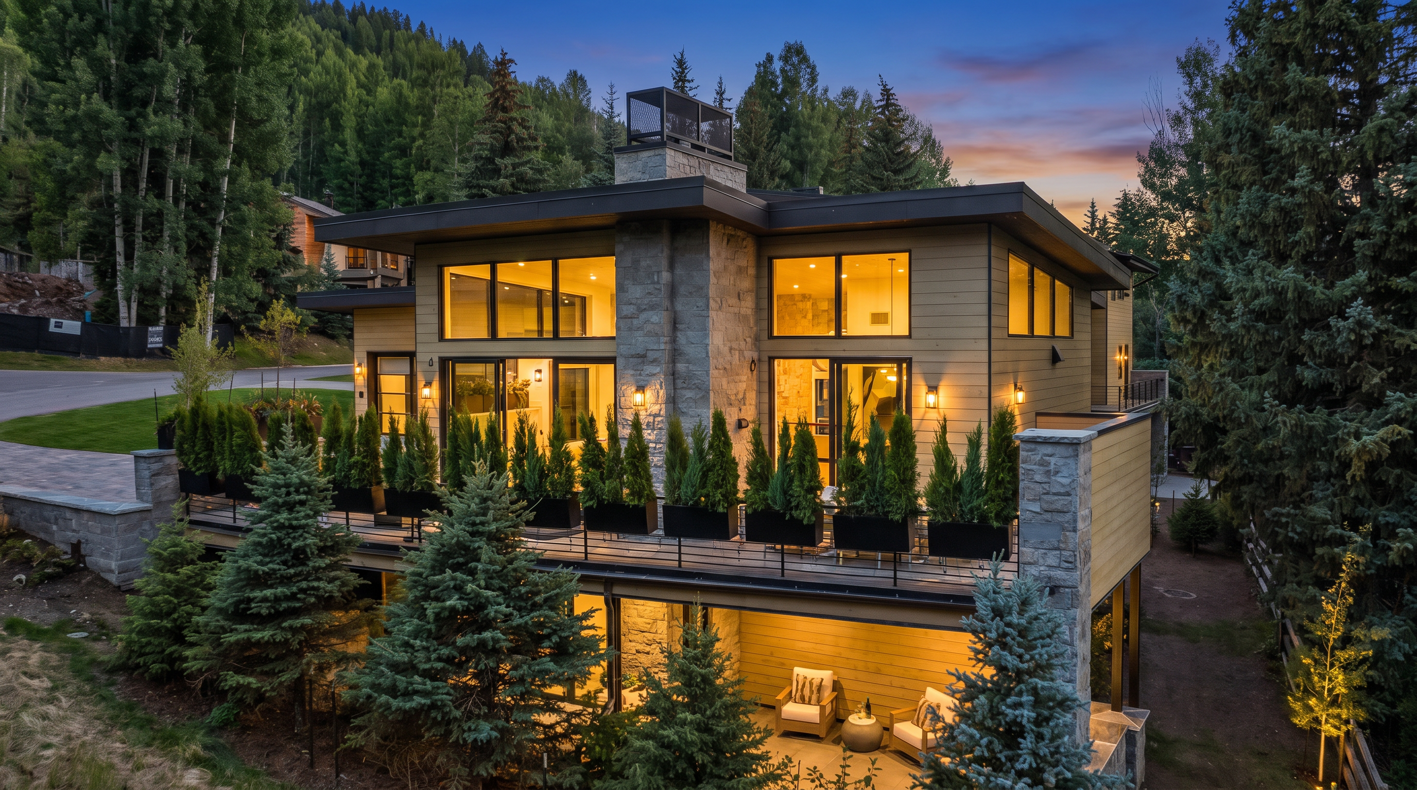 This stunning contemporary residence showcases a blend of natural stone and wood siding, set against a lush, forested mountain backdrop. The house features floor-to-ceiling windows and a spacious wrap-around deck adorned with orderly potted evergreens, creating a seamless transition between indoor and outdoor living. With its warm evening lighting and sophisticated modern design, the property exudes an inviting and luxurious appeal.