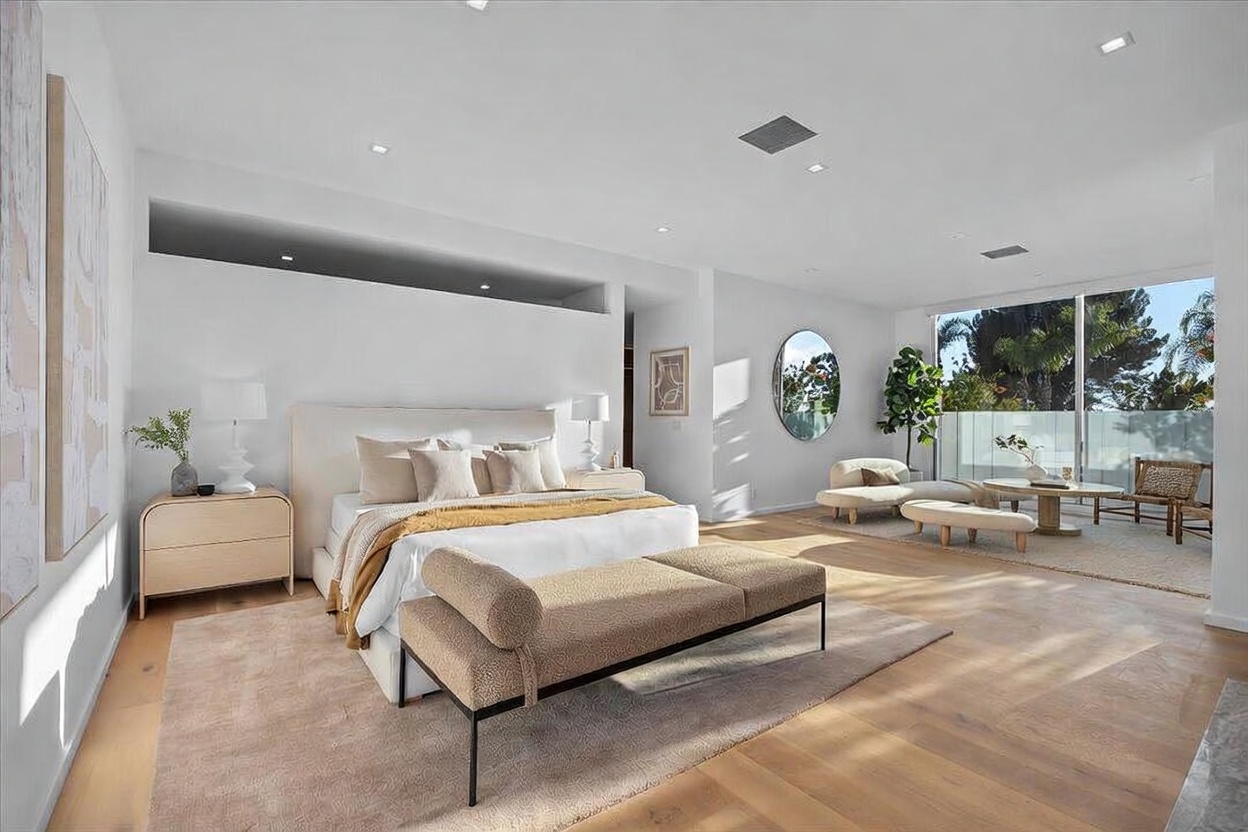 This spacious primary bedroom features a neutral color palette, light wood flooring, and a large area rug that anchors the bed. The room includes a modern upholstered bed frame, a sleek bench at the foot of the bed, and a separate seating area near the large glass sliding doors that lead to a balcony. A circular mirror and minimalist decor enhance the bright, airy atmosphere of this contemporary suite.