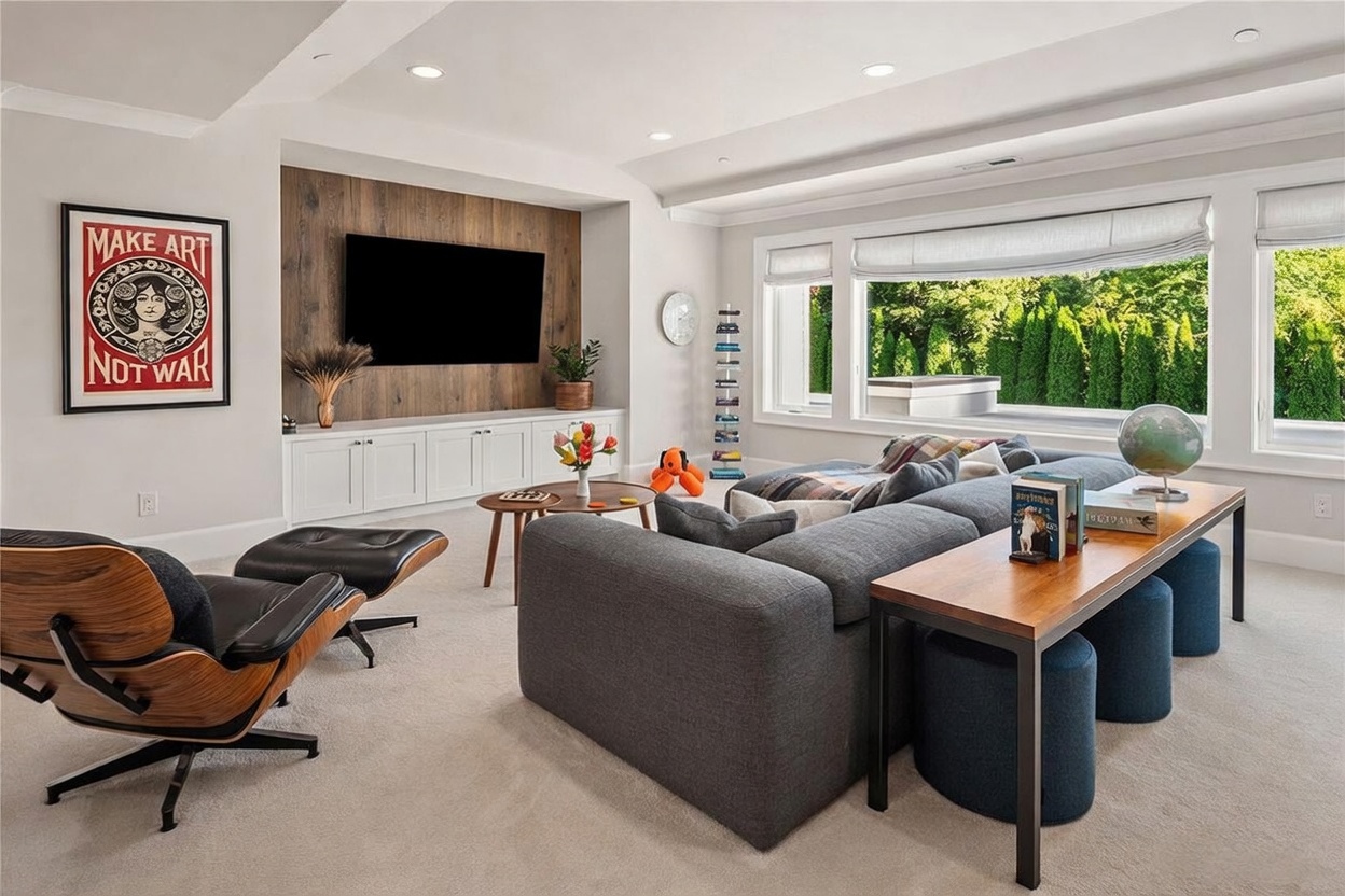 This spacious living room features a large, comfortable gray sectional sofa positioned to face a media wall with a wood-paneled accent and built-in white cabinetry. A classic lounge chair with an ottoman sits to the left, while a console table behind the sofa provides additional seating with dark blue stools. Large windows along the back wall offer a bright, scenic view of the lush greenery outside, creating an inviting and relaxed atmosphere.