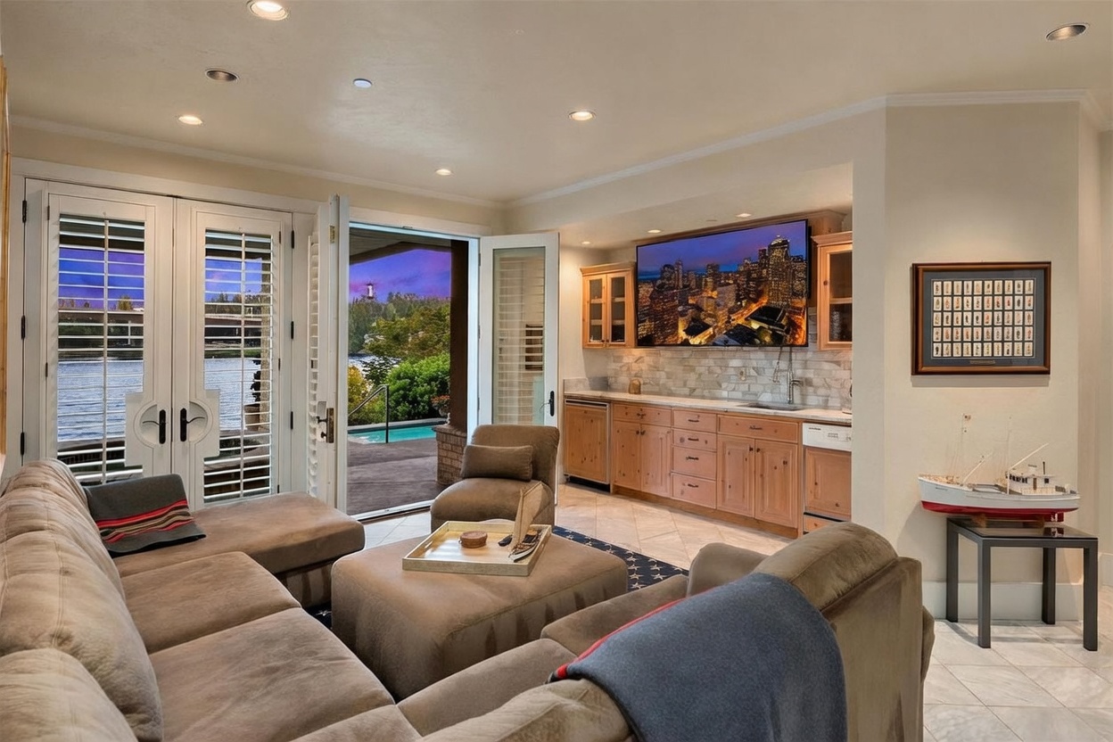 This inviting living room features a comfortable sectional sofa and a central ottoman, creating a cozy space for relaxation. A wet bar with light wood cabinetry and a stone backsplash is integrated into the wall, topped by a large television displaying a vibrant city skyline. The room opens through double glass doors to a scenic outdoor patio area with a pool, offering a seamless transition between indoor and outdoor living.