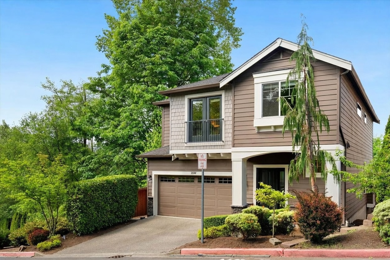 This two-story suburban home features a blend of horizontal siding and shingle-style accents, complemented by a two-car garage and a small balcony on the upper level. The front yard is landscaped with manicured hedges and shrubs, while a paved driveway leads to the garage entrance. The perspective is a straight-on eye-level shot, capturing the home's inviting curb appeal against a clear blue sky.