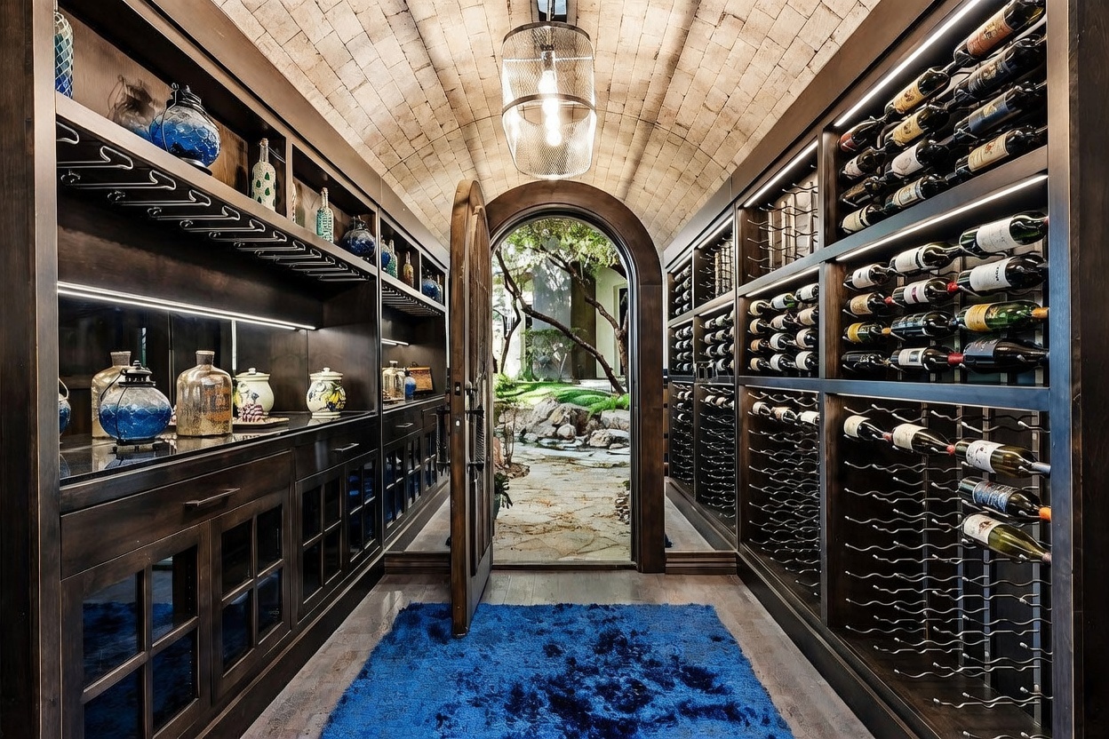 This sophisticated wine cellar features a barrel-vaulted ceiling with textured masonry and custom dark wood cabinetry for extensive bottle storage. A central arched doorway opens to a lush, sunlit garden view, creating a seamless transition between the climate-controlled interior and the outdoors. The space is accented by a vibrant blue rug and decorative glass vessels, offering a luxurious and atmospheric aesthetic.