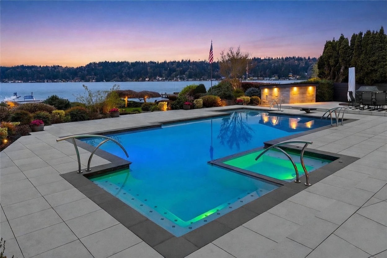 This stunning outdoor space features a modern, rectangular swimming pool with an integrated spa, both illuminated with vibrant green and blue lighting. The pool deck is paved with large, light-colored stone tiles, and the area overlooks a serene lake at sunset, creating a luxurious and tranquil atmosphere. The composition captures the elegant transition from the manicured landscaping to the expansive water view in the background.