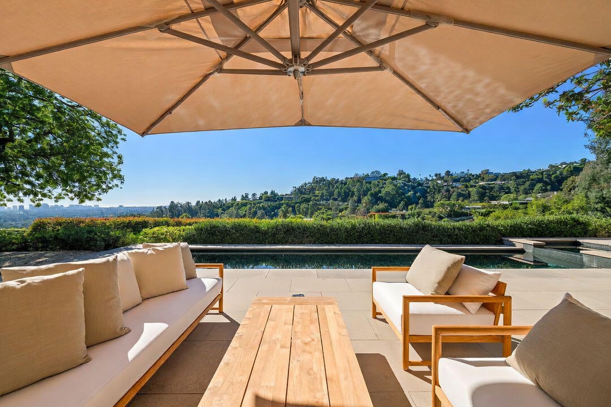 This luxurious outdoor patio space features a modern teak wood coffee table and comfortable lounge seating arranged under a large, neutral-toned cantilever umbrella. The area overlooks a sleek, rectangular infinity-edge pool that seamlessly blends into the lush, rolling hillside landscape beyond. The scene captures a serene and sophisticated atmosphere, perfect for high-end outdoor entertaining with panoramic views.