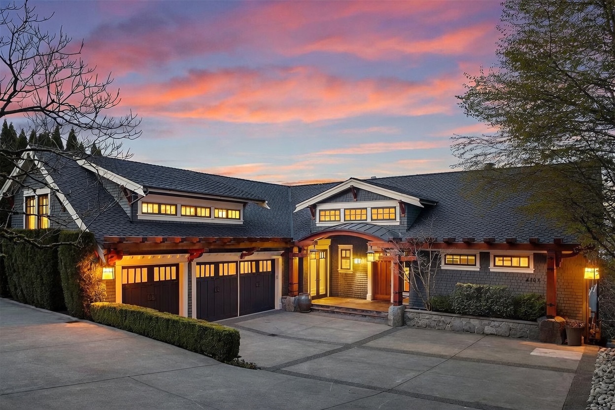 This stunning craftsman-style home is captured at dusk, showcasing a warm, inviting facade with dark shingle siding and natural wood accents. The perspective highlights the expansive driveway leading to a multi-car garage and a welcoming, illuminated entryway under a curved roofline. The vibrant sunset sky provides a dramatic backdrop, emphasizing the home's sophisticated architectural details and curb appeal.
