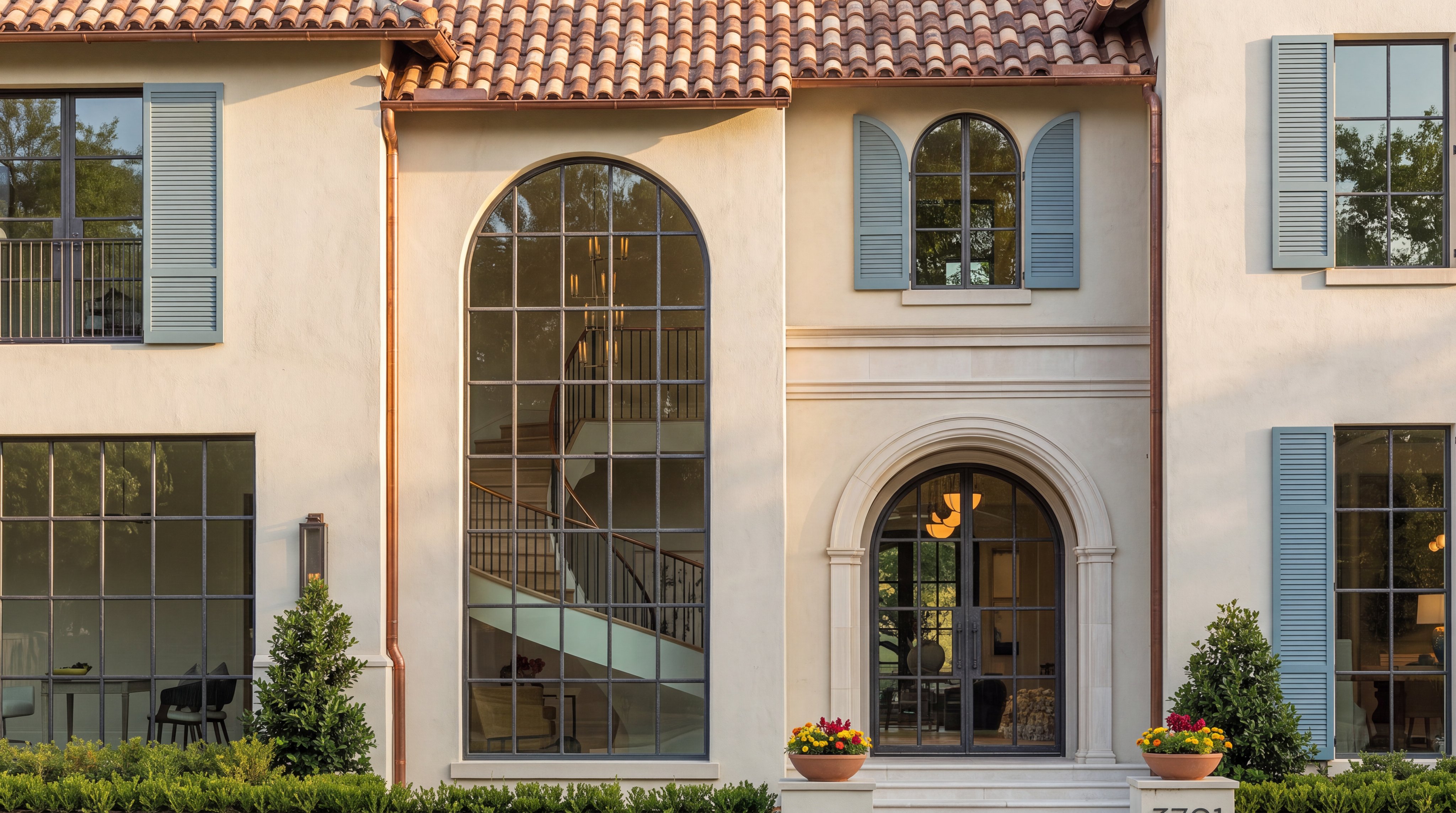 This elegant home features a European-inspired aesthetic with a light stucco facade, terracotta tile roof, and distinct arched windows and doorways. The symmetry of the blue shutters, copper gutters, and manicured landscaping creates a sophisticated and welcoming curb appeal. A grand, multi-story arched window reveals a glimpse of an interior staircase, conveying luxury and architectural character.