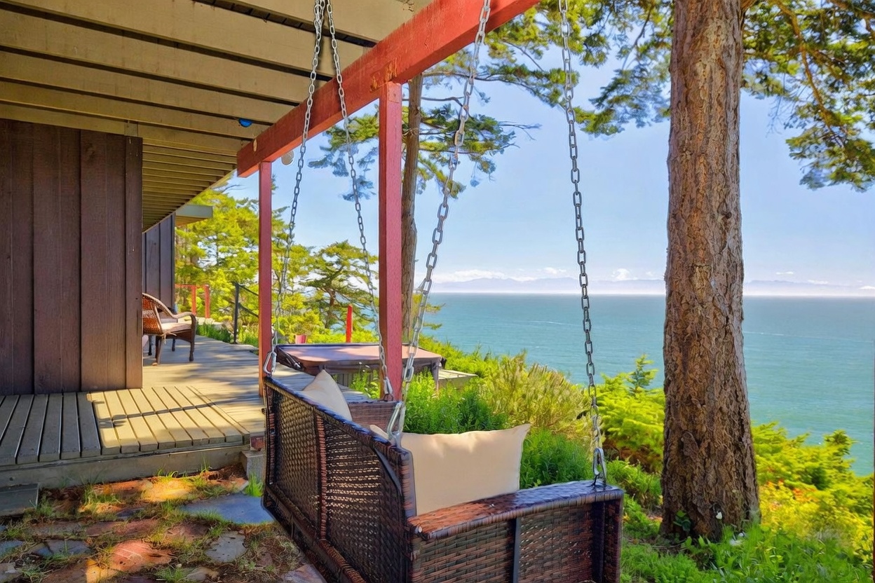 This inviting outdoor deck features a hanging wicker swing with plush cushions, providing a perfect vantage point for the expansive ocean view. The wooden deck extends toward a hot tub nestled near the edge, framed by lush greenery and a tall pine tree. The scene captures a serene, coastal atmosphere with a clear view of distant mountains across the water.