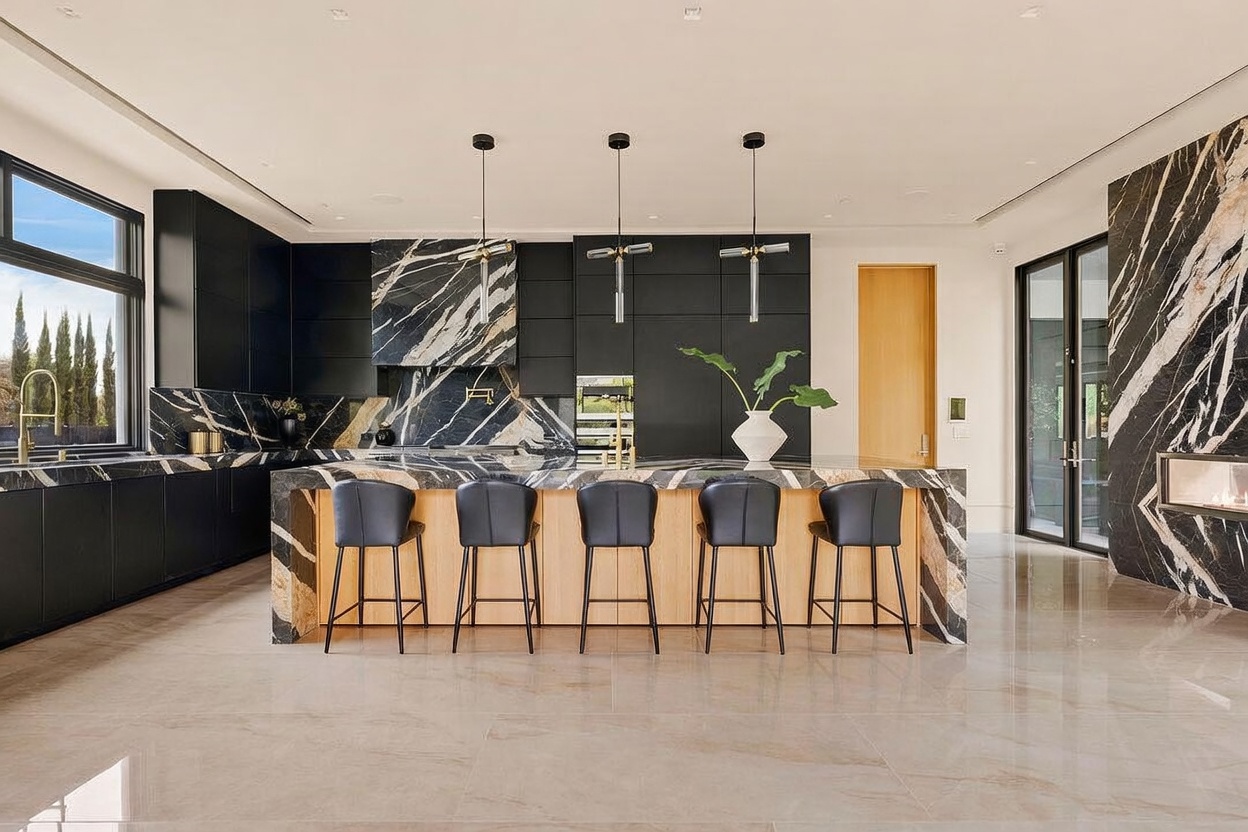 This sophisticated, modern kitchen features a striking contrast between dark, matte cabinetry and bold, veined stone surfaces on the island and backsplash. A large central island provides ample seating with five dark stools, illuminated by three sleek, minimalist pendant lights. The space is anchored by light-toned, reflective flooring and a warm wood-paneled wall, creating a luxurious and high-end aesthetic.