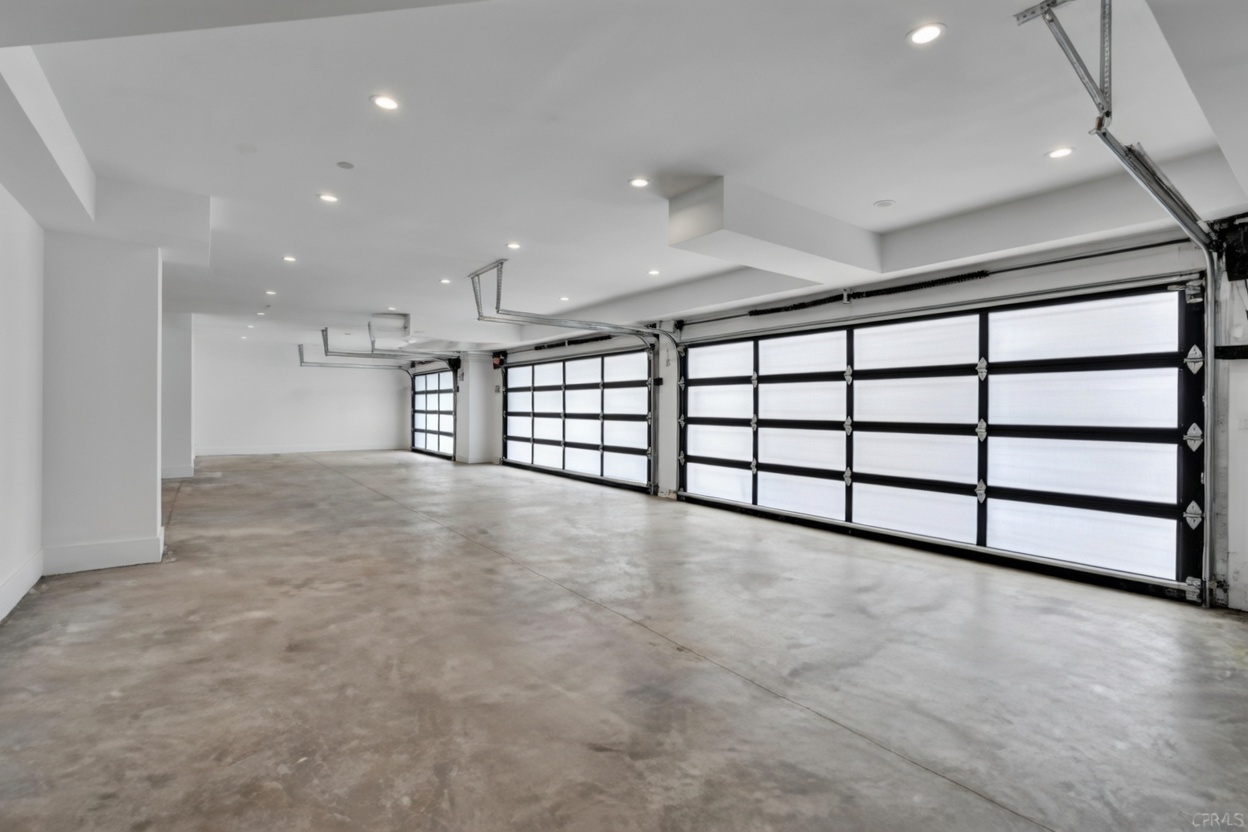 This spacious, multi-car garage features a clean, minimalist aesthetic with smooth concrete flooring and bright recessed lighting. The space is defined by several large, modern glass-paneled garage doors that allow for significant natural light, creating an airy and expansive atmosphere. The perspective is wide-angle, showcasing the depth and capacity of the garage, which appears to be a high-end, contemporary design.