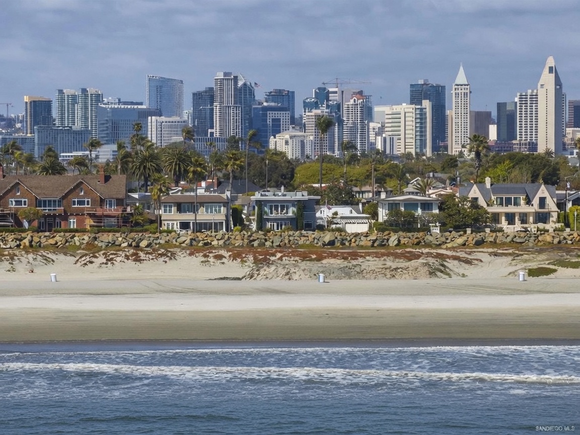 This wide-angle, eye-level shot captures a scenic coastal landscape featuring a sandy beach in the foreground and a row of residential homes nestled behind a rocky seawall. Beyond the homes, a lush green belt transitions into the impressive, dense skyline of a major city, showcasing a mix of modern high-rise architecture and iconic towers. The perspective emphasizes the unique proximity of luxury waterfront living to a vibrant urban center under a bright, partly cloudy sky.