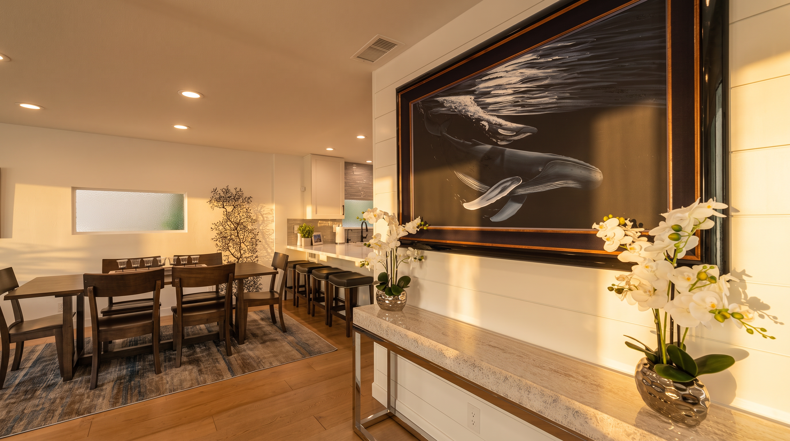 This modern dining area features a dark wood table and chairs set upon a textured area rug, creating a sophisticated contrast against the light hardwood floors. An adjacent kitchen breakfast bar with stools provides additional seating, while the wall is adorned with a large, striking whale painting and a decorative white orchid arrangement. The space is bright and airy, blending elegant decor with functional living areas for a cohesive open-concept layout.