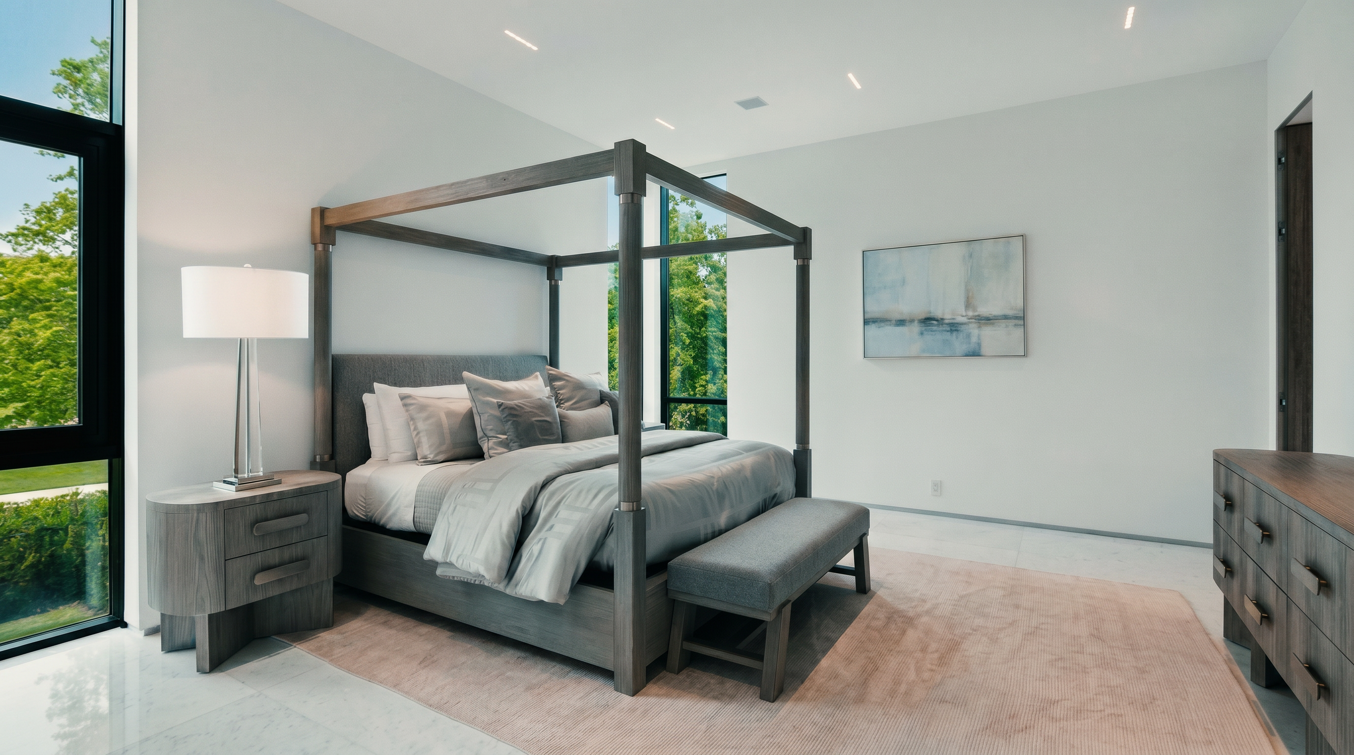 This spacious primary bedroom features a modern four-poster bed as the central focal point, complemented by a matching nightstand and a long dresser. The room is bathed in natural light from large windows that offer views of the surrounding greenery, while a neutral color palette and clean lines create a serene atmosphere. A soft area rug sits beneath the bed and bench, adding texture to the polished light-colored flooring.