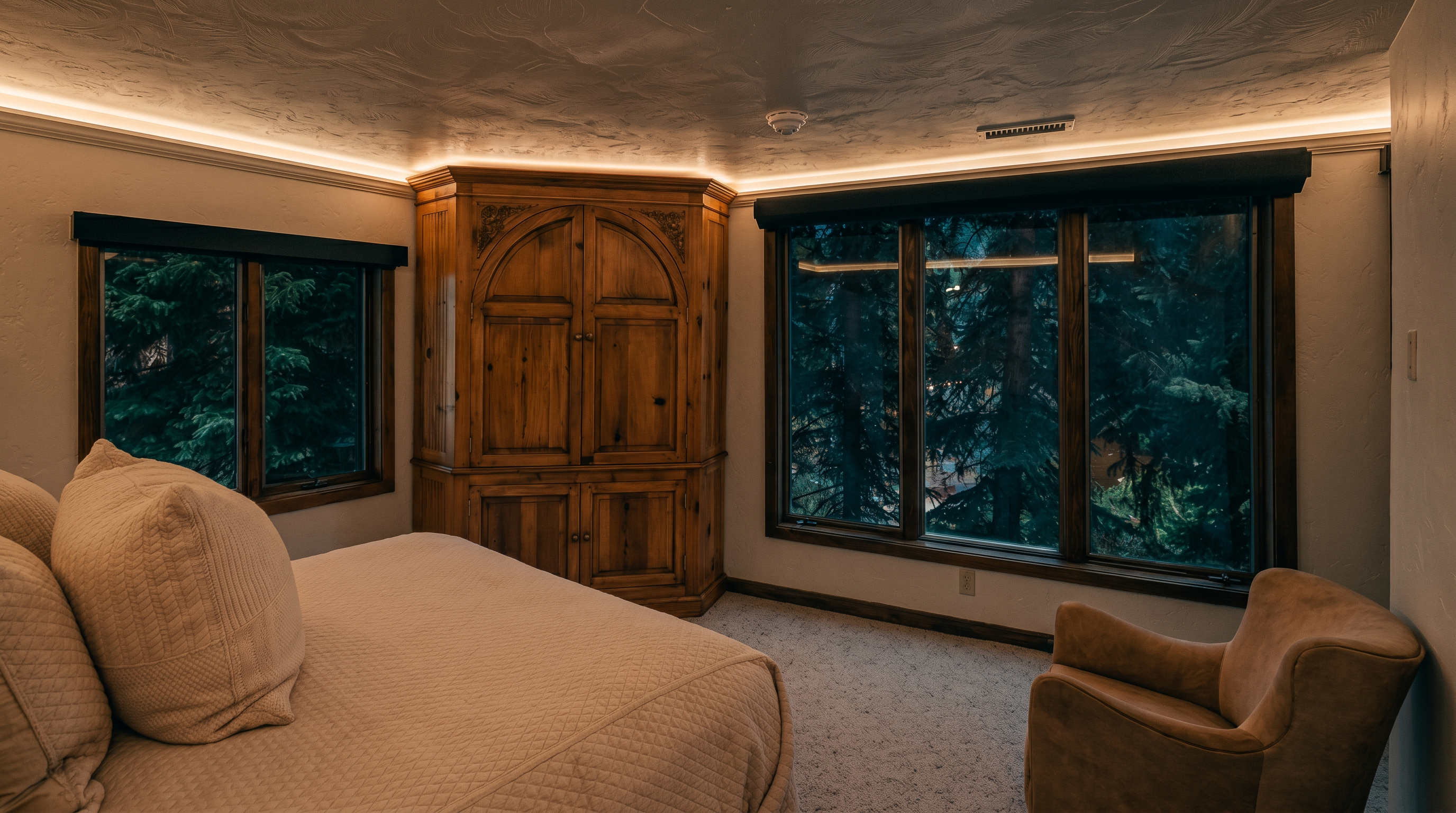 This bedroom features a neutral color palette with a large bed topped with a textured quilt and a plush armchair positioned near a set of large windows. A substantial wooden armoire with arched detailing sits in the corner, providing a classic focal point against the light-colored walls. The room is bathed in natural light, offering a serene view of the surrounding forest through the windows.