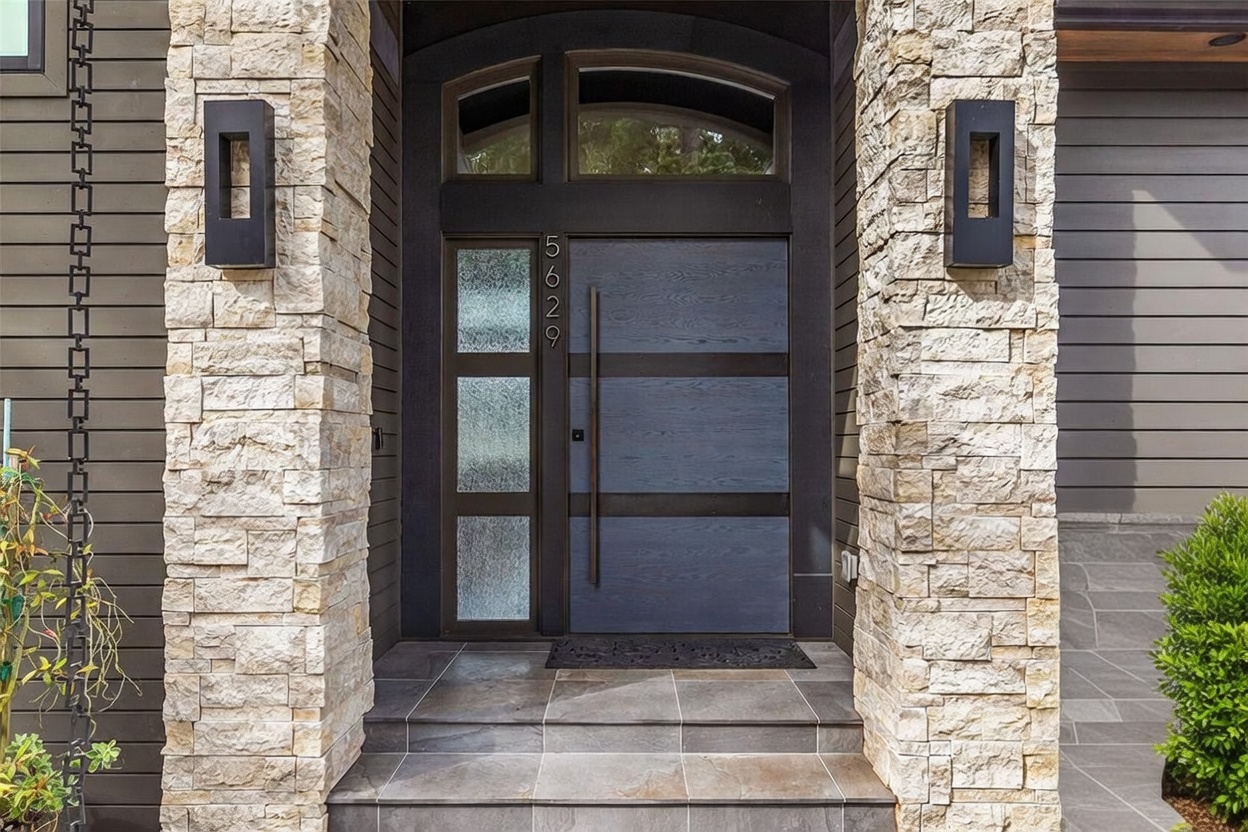 This modern entryway features a striking front door with horizontal wood panels and a vertical glass sidelight displaying the house number 5629. The entrance is framed by two substantial stone-clad pillars, each accented with a sleek, rectangular black wall sconce. The perspective is a direct, eye-level shot that highlights the clean lines, textured stone, and contemporary architectural design of the home's facade.