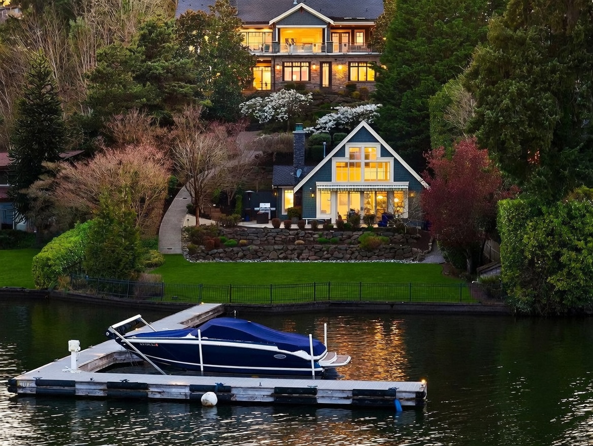 This stunning waterfront property features a multi-level landscape with a charming cottage-style home nestled near the water and a larger, grand residence situated on the hill above. The scene is captured from a low-angle perspective across the water, showcasing a private dock with a boat, a manicured lawn, and tiered stone retaining walls. The warm glow from the interior lights creates an inviting and serene atmosphere, highlighting the architectural beauty of both homes against the lush, wooded backdrop.