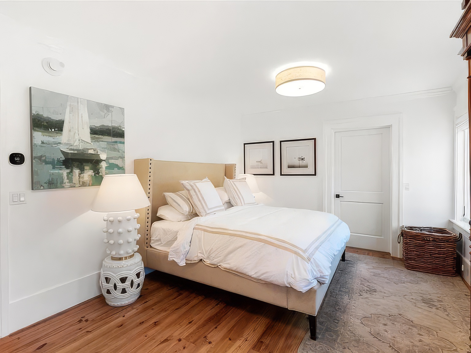 This bright and airy bedroom features a neutral color palette with a beige upholstered headboard and white bedding accented by striped pillows. A unique, textured white lamp sits on a matching ceramic base, while a sailboat painting adds a nautical touch to the wall. The room is completed by warm wood flooring, a patterned area rug, and a woven basket, creating a serene and inviting atmosphere.