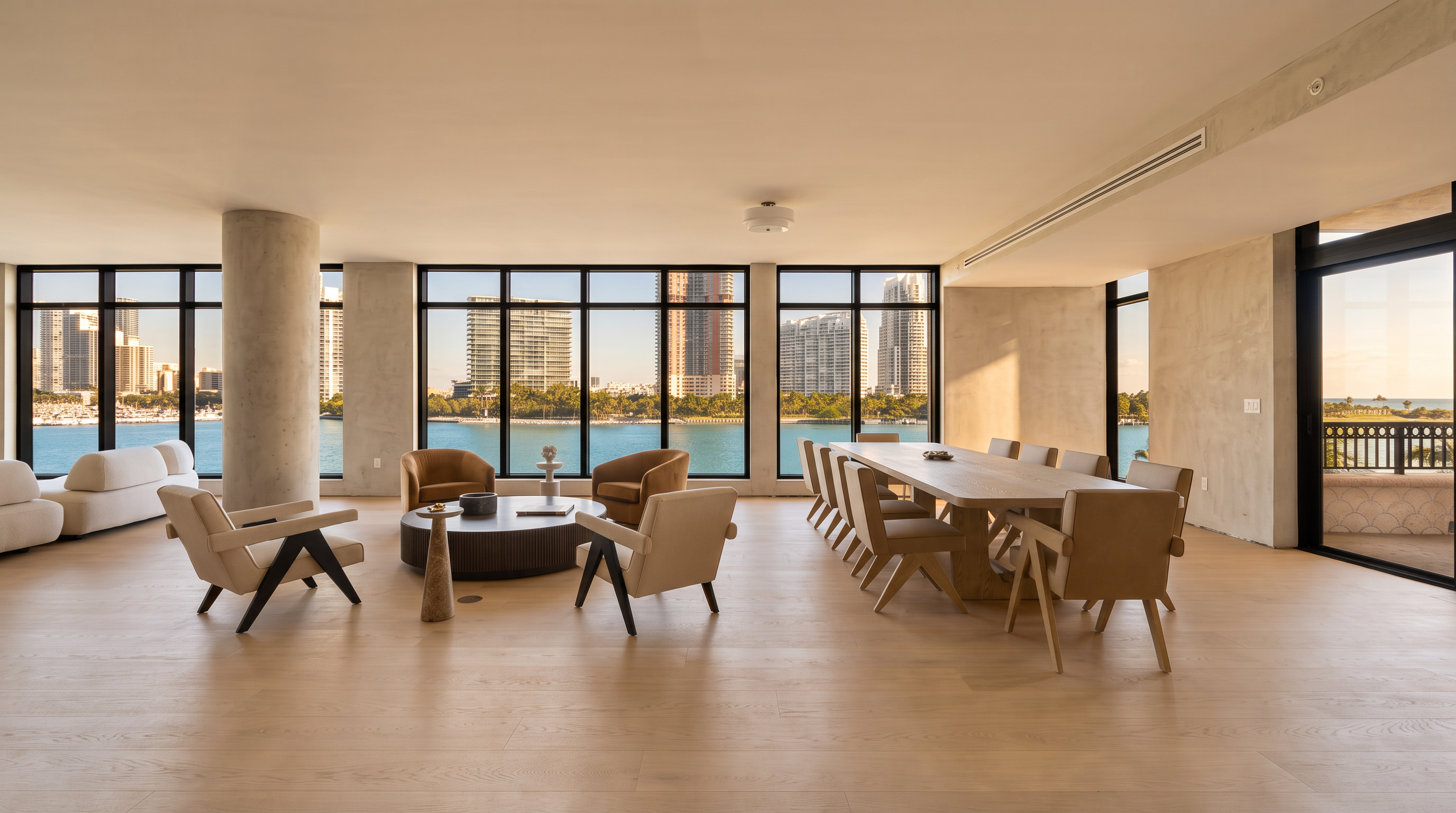 This expansive, open-concept living and dining area features light wood flooring and a neutral color palette that emphasizes the bright, airy atmosphere. Large floor-to-ceiling windows span the entire back wall, offering panoramic views of the water and city skyline. The space is furnished with a contemporary seating area and a long dining table, creating a sophisticated and seamless transition between indoor living and the scenic outdoor backdrop.