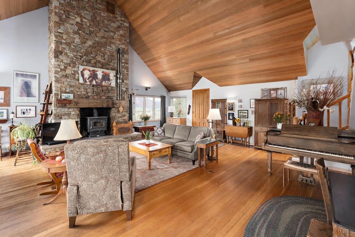 This spacious living room features a vaulted ceiling with warm wood paneling and a prominent stone-clad chimney breast that serves as the focal point. The room is furnished with a comfortable sofa, a patterned armchair, and a wooden coffee table, all arranged on a large area rug over hardwood floors. A piano sits to the right, and the space is filled with natural light from windows that offer a view of the landscape beyond.