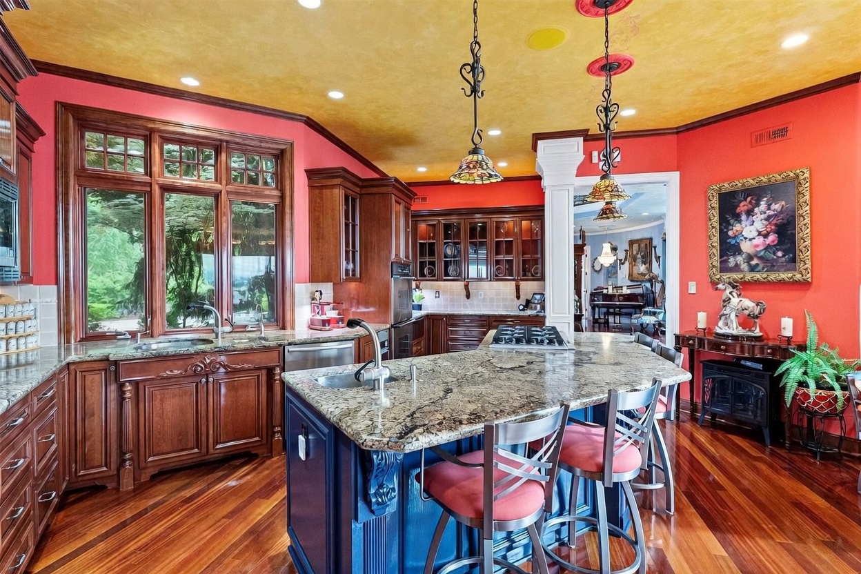 This vibrant kitchen features a bold design with bright red walls, a textured yellow ceiling, and rich wood cabinetry. A large central island with a stone countertop and a blue base provides ample workspace and seating, complemented by decorative pendant lighting. The space is well-lit by recessed lighting and large windows that overlook the exterior greenery, creating a warm and eclectic atmosphere.