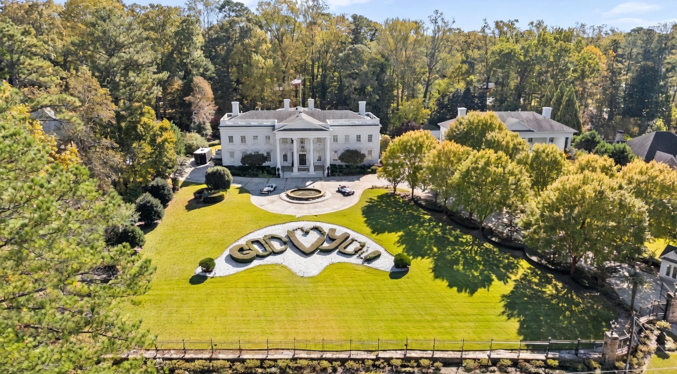 This high-angle aerial shot captures a grand, white neoclassical mansion set against a backdrop of dense, mature trees. The property features a prominent circular driveway with a central fountain, a sprawling front lawn adorned with a large, decorative 'GOD <3 YOU' landscaping feature, and a secondary structure to the right. The perspective emphasizes the estate's significant scale, symmetrical architecture, and expansive, manicured grounds.