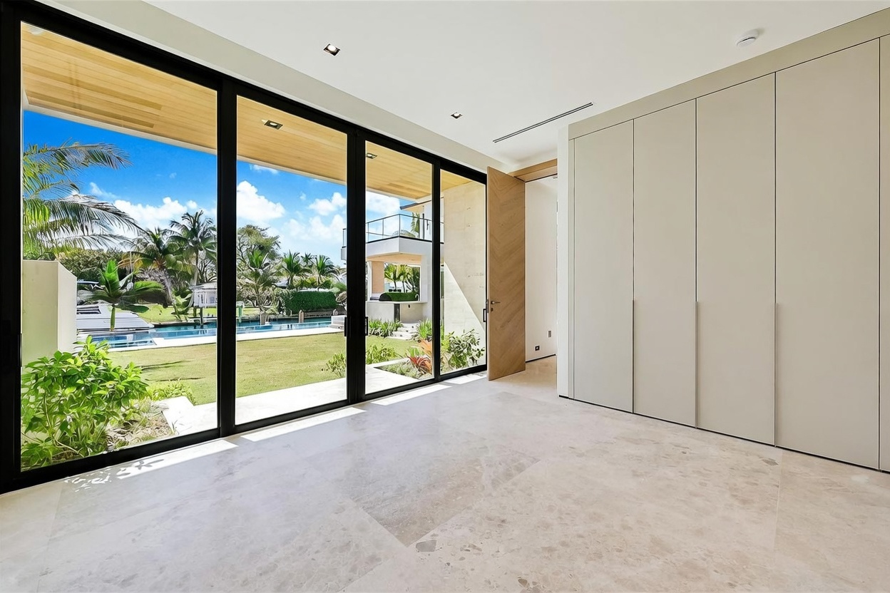 This bright, minimalist room features large floor-to-ceiling glass doors that open to a lush backyard and pool area. The space is finished with light-toned stone flooring and a wall of sleek, handle-less cabinetry, creating a seamless and modern aesthetic. A wooden door with a chevron pattern adds a touch of warmth to the neutral interior, while recessed lighting maintains a clean, uncluttered look.