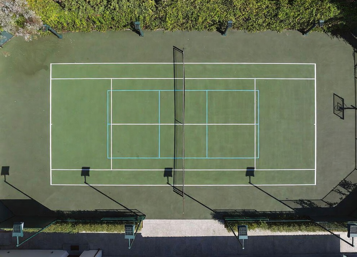 This high-angle aerial view captures a well-maintained tennis court surrounded by lush green hedges. The court features a central net, white boundary lines, and a basketball hoop positioned at one end, set against a backdrop of mature trees. The perspective provides a clear, symmetrical look at the recreational space, highlighting its private and secluded setting.