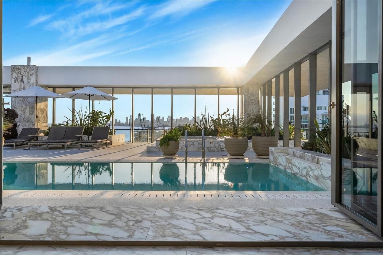 This luxurious rooftop terrace features a private swimming pool framed by elegant stone tiling and large glass panels that offer an expansive view of the city skyline. The space is furnished with lounge chairs under umbrellas and decorative potted plants, creating a serene outdoor retreat. The perspective is captured from an indoor threshold, looking out toward the sun-drenched pool deck and the distant urban horizon.