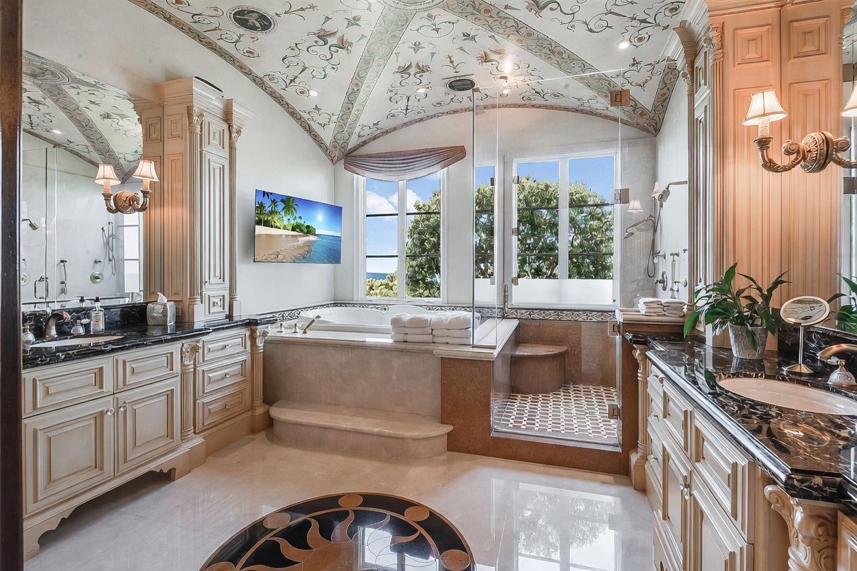 This luxurious primary bathroom features dual vanities with dark stone countertops and light-toned cabinetry, flanking a central soaking tub and a glass-enclosed shower. The space is highlighted by an ornate, hand-painted vaulted ceiling and large windows that provide natural light and a view of the outdoors. A flat-screen television is mounted on the wall above the tub, contributing to the high-end, spa-like atmosphere of the room.