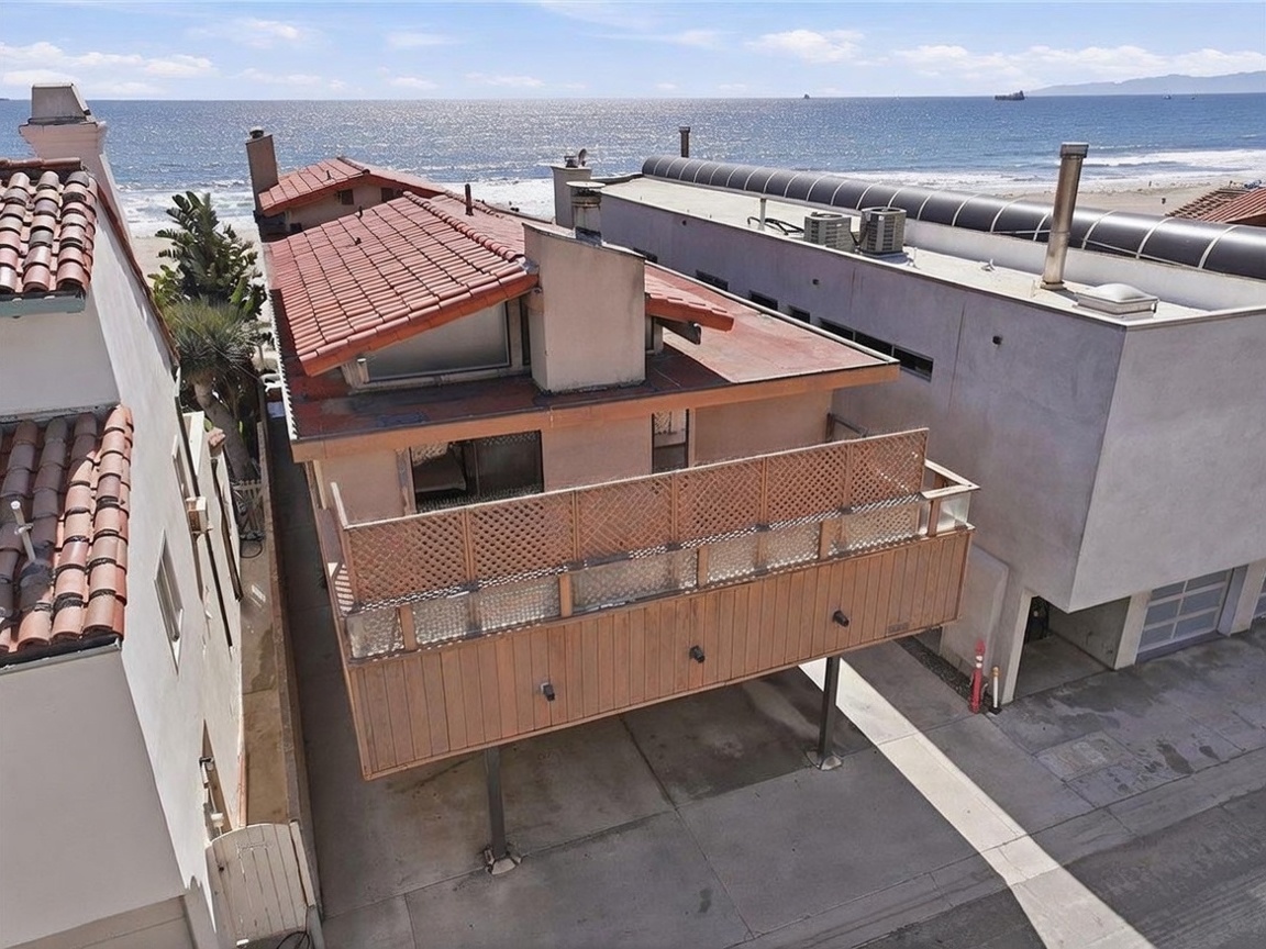 This high-angle aerial shot captures a beachside property with a distinctive elevated wooden deck supported by metal pillars. The house features a terracotta-tiled roof and a prominent chimney stack, situated directly adjacent to the sandy shoreline and the ocean. The perspective highlights the home's unique positioning and its immediate proximity to the coastal environment.