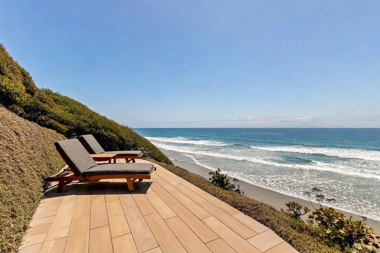 This elevated wooden deck offers a serene, panoramic view of the ocean and sandy coastline. Two lounge chairs with dark cushions are positioned to take advantage of the coastal scenery, set against a backdrop of lush hillside greenery. The perspective is wide and cinematic, capturing the tranquil atmosphere of a private seaside retreat.