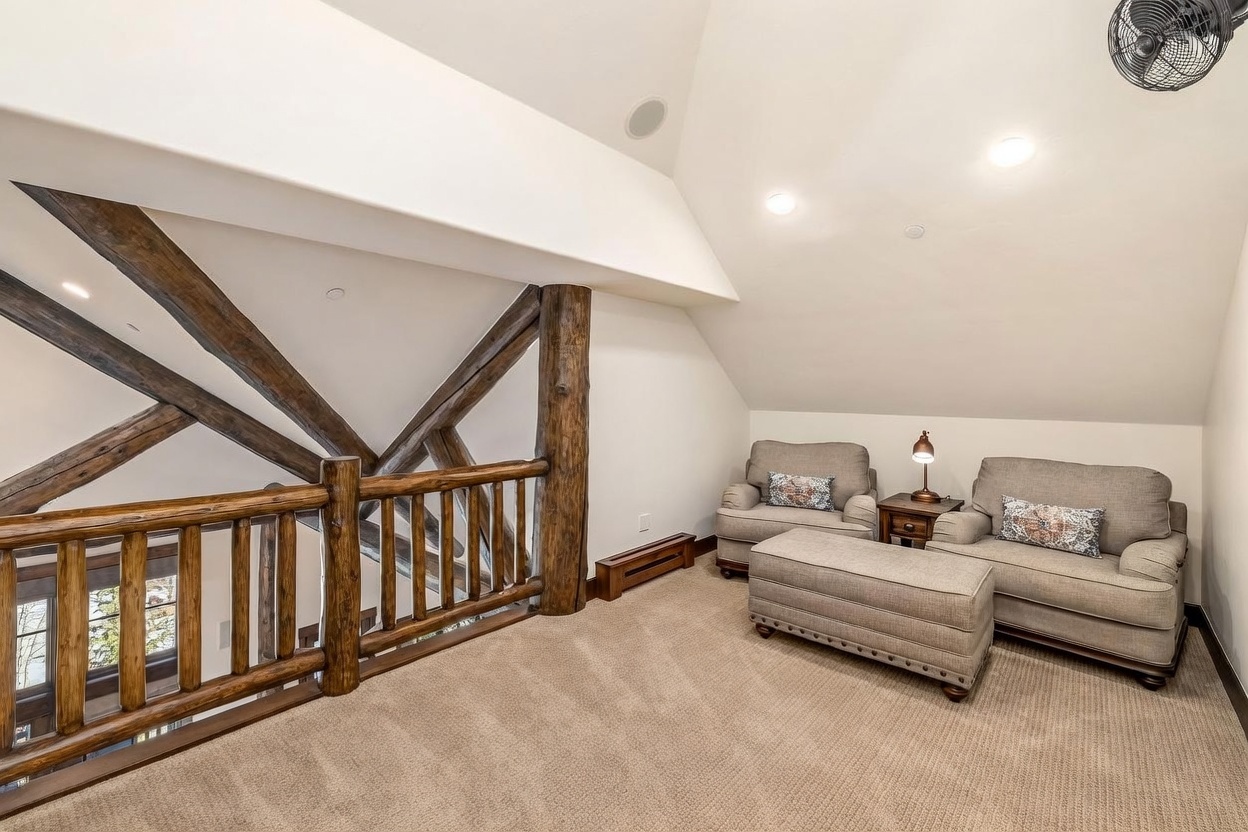 This cozy loft area features a rustic aesthetic with exposed timber beams and a wooden railing overlooking the level below. The space is furnished with two comfortable armchairs, a matching ottoman, and a small side table topped with a lamp, creating a quiet reading nook under the sloped ceiling. The neutral carpet and soft lighting contribute to a warm, inviting atmosphere perfect for relaxation.
