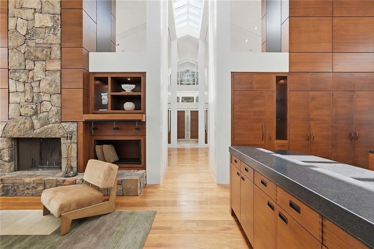This spacious living area features a prominent stone-clad chimney breast and warm wood cabinetry that extends across the wall. A comfortable armchair sits on a textured rug near the hearth, while a long kitchen island with a dark countertop occupies the foreground. The perspective looks down a central hallway toward a bright, light-filled entryway with a high, vaulted ceiling and skylights.