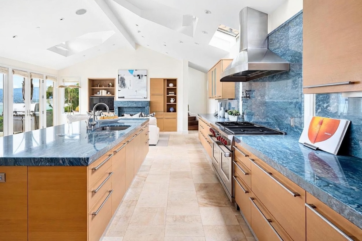 This spacious, open-concept kitchen features a large central island and extensive perimeter cabinetry finished in light wood tones. The space is defined by striking blue stone countertops and a matching backsplash, complemented by stainless steel appliances and a professional-grade range hood. Large skylights and glass doors allow natural light to flood the room, creating an airy atmosphere that flows seamlessly into the adjacent living area.