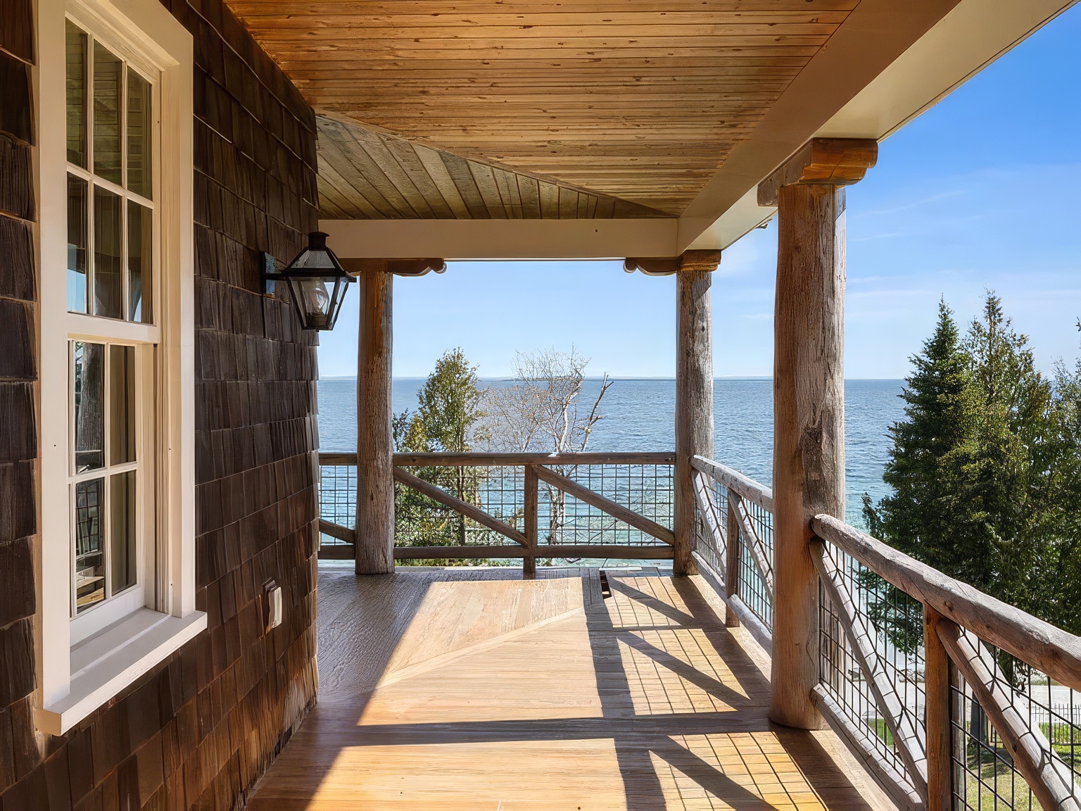 This covered deck offers a serene, elevated view of the water, framed by rustic log support columns and a wooden ceiling. The space features a natural wood deck floor and a railing constructed from logs and wire mesh, blending seamlessly with the surrounding coastal landscape. The perspective looks outward from the porch, highlighting the tranquil connection between the home's architecture and the expansive lake or ocean view.