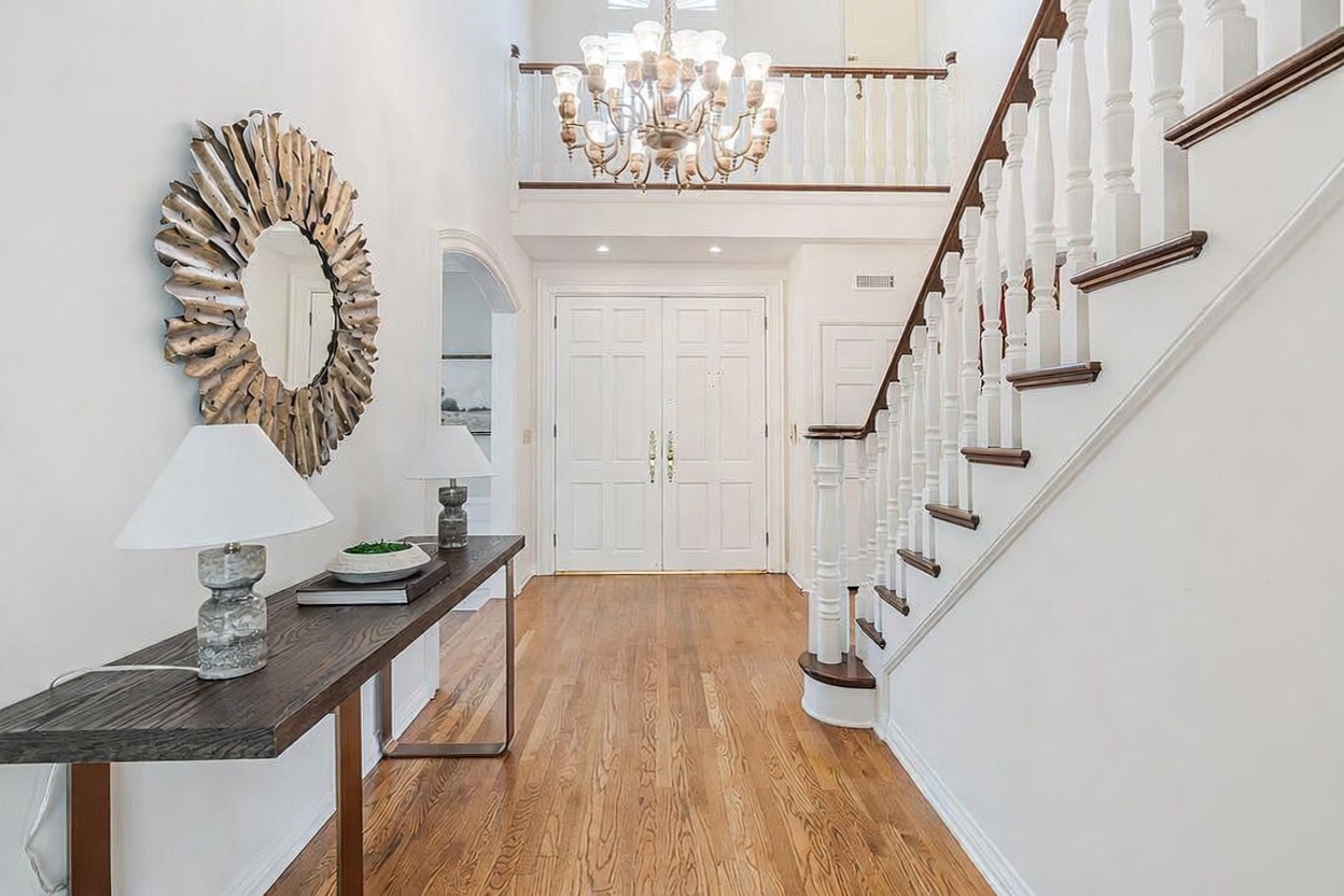 This grand entryway features a high-ceiling foyer with a prominent staircase and a decorative chandelier hanging from the upper level. A long console table sits against the wall, topped with a lamp and a large, textured sunburst-style mirror. The space is finished with light wood flooring and white trim, creating a bright and welcoming atmosphere.