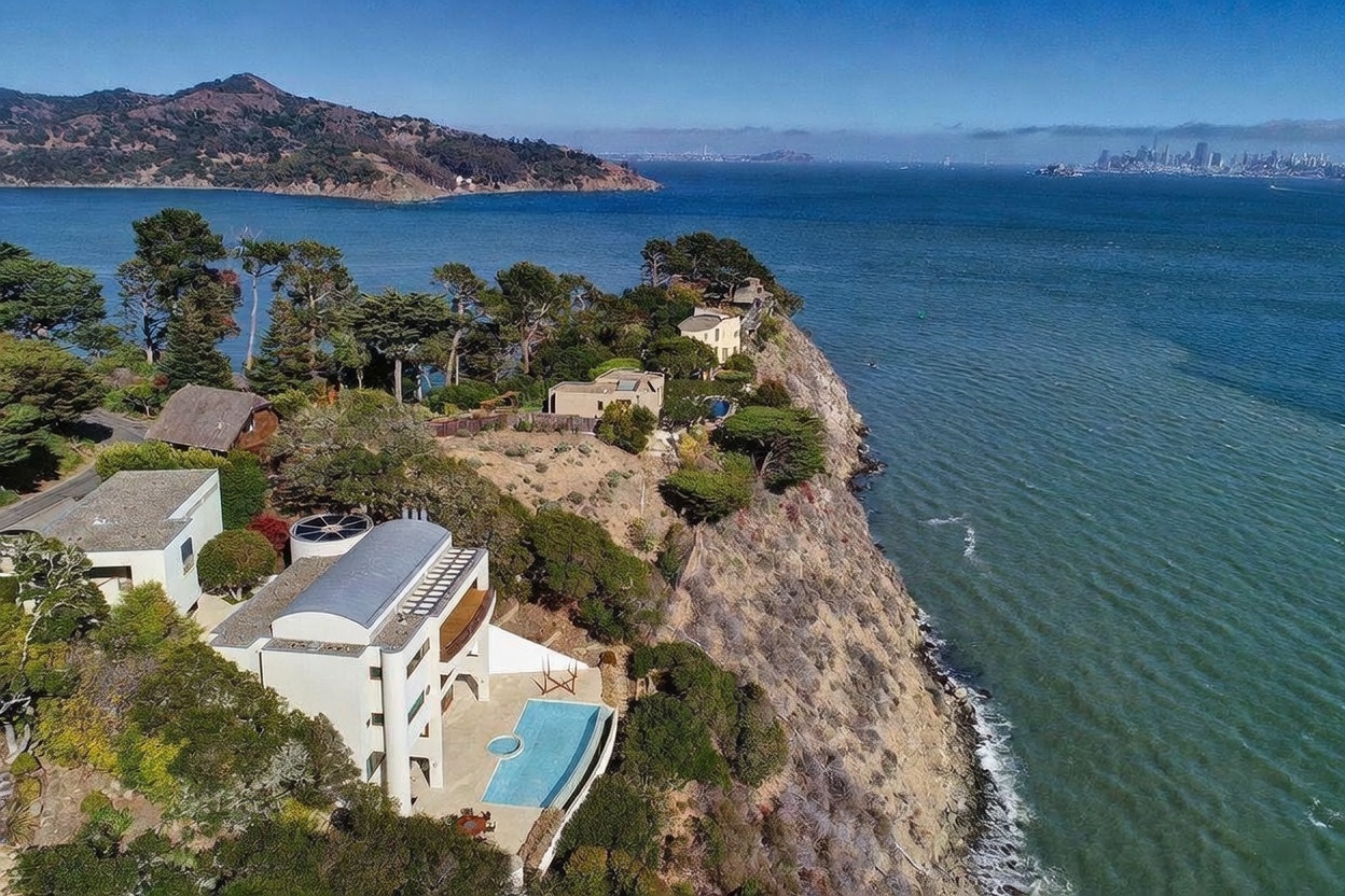 This high-angle aerial view captures a stunning coastal property perched on a rugged cliffside overlooking the bay. The modern, multi-level residence features a distinctive curved roofline and a private swimming pool that appears to blend into the horizon. The surrounding landscape is characterized by mature trees and natural terrain, with a panoramic view of the distant city skyline across the water.