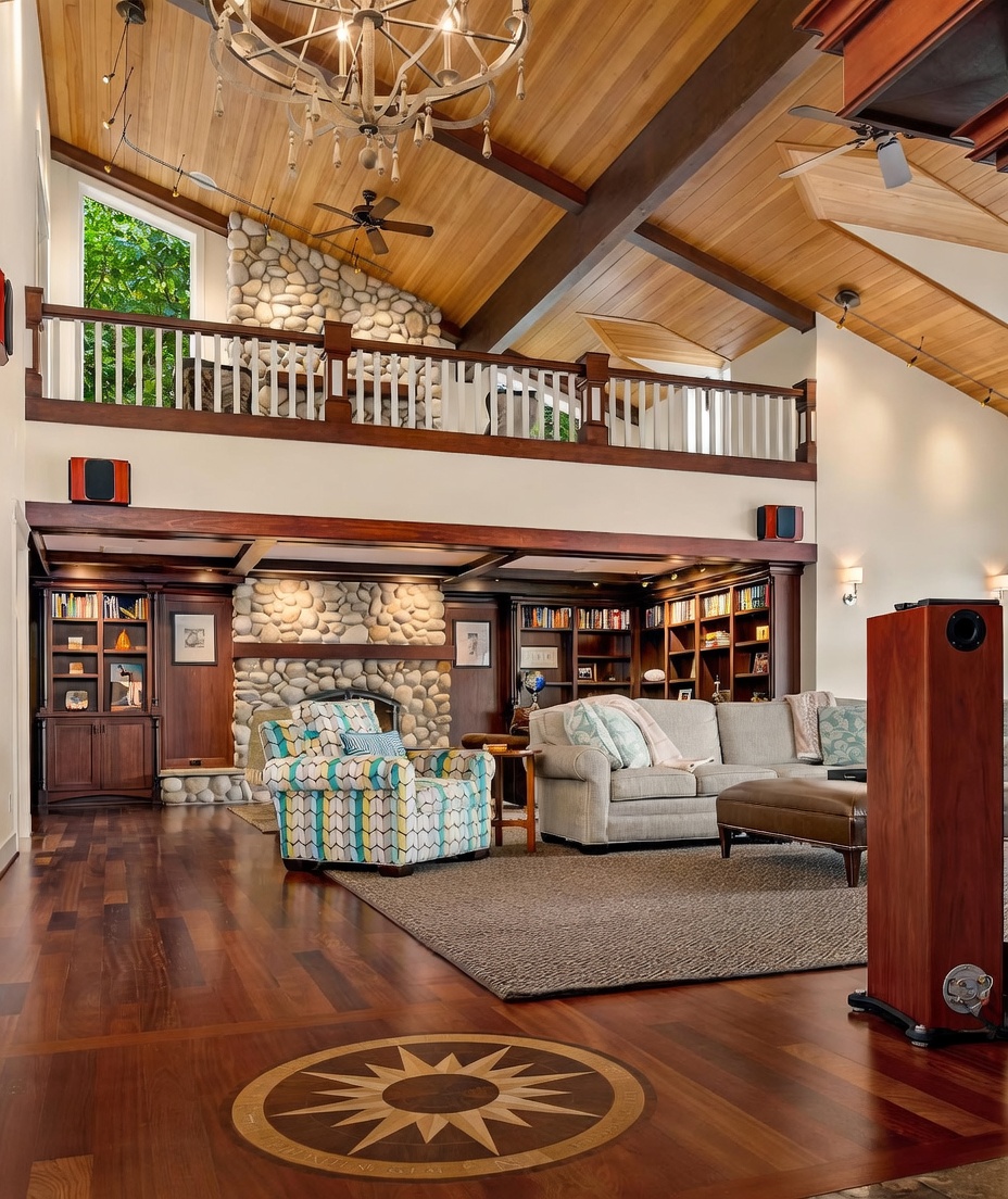 This expansive living room features a vaulted wood-paneled ceiling with exposed beams and a prominent stone-clad chimney breast that anchors the space. The room includes a cozy seating area with a patterned armchair and a neutral sofa, set upon a large area rug over rich-toned hardwood floors with a decorative inlay. A lofted balcony with a white railing overlooks the main living area, creating an open and airy atmosphere with a rustic, lodge-inspired aesthetic.