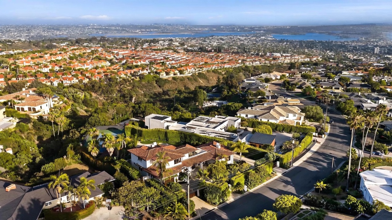 This high-angle aerial view captures a sprawling residential neighborhood in a coastal setting, featuring a prominent multi-level home with a terracotta-tiled roof in the foreground. The property is surrounded by lush landscaping, mature palm trees, and a private driveway, with a vast suburban landscape and a distant bay visible in the background. The perspective highlights the home's integration into the hillside terrain and its proximity to the surrounding community.