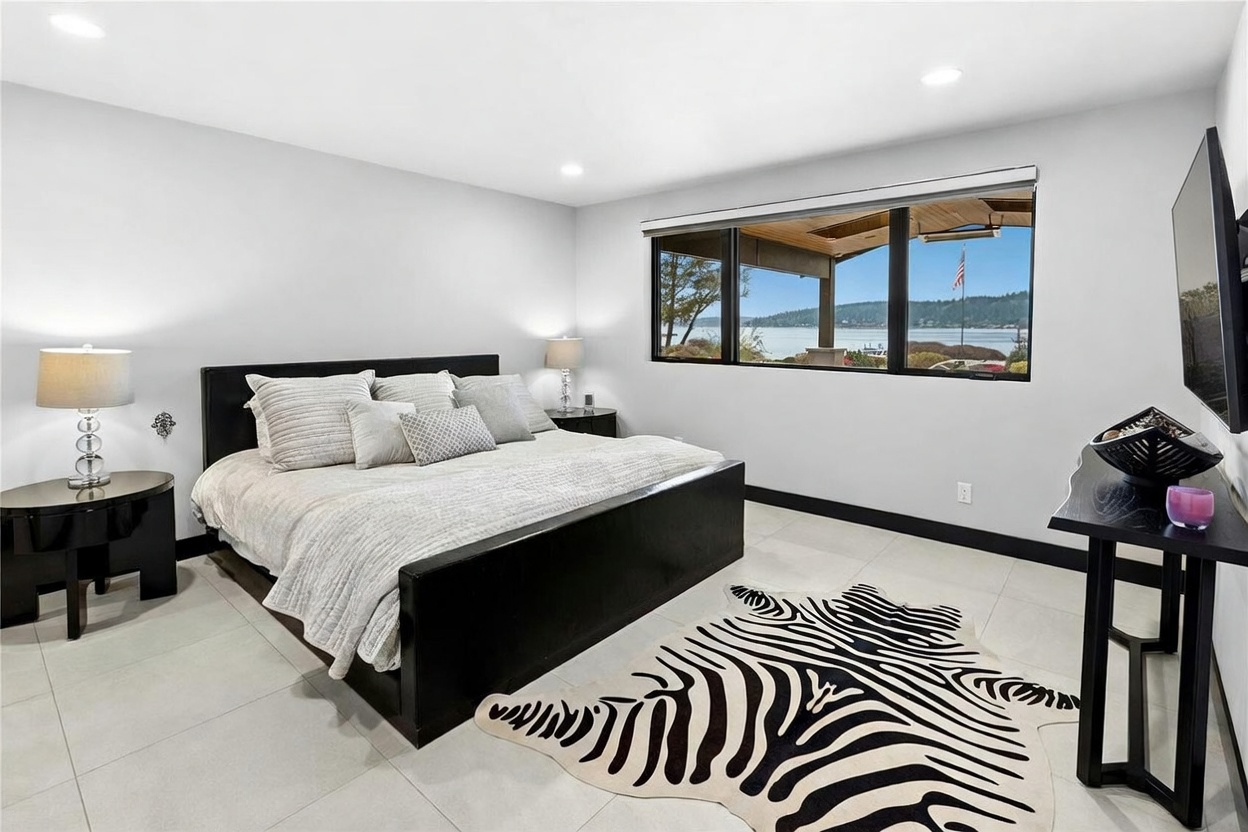 This primary bedroom features a contemporary design with a large, dark-framed bed as the centerpiece, complemented by a striking zebra-print area rug on light-colored tile flooring. A wide window provides a scenic, panoramic view of the water and outdoor landscape, creating a serene and luxurious atmosphere. The room is minimalist and bright, with sleek bedside tables and lamps that enhance the modern aesthetic.