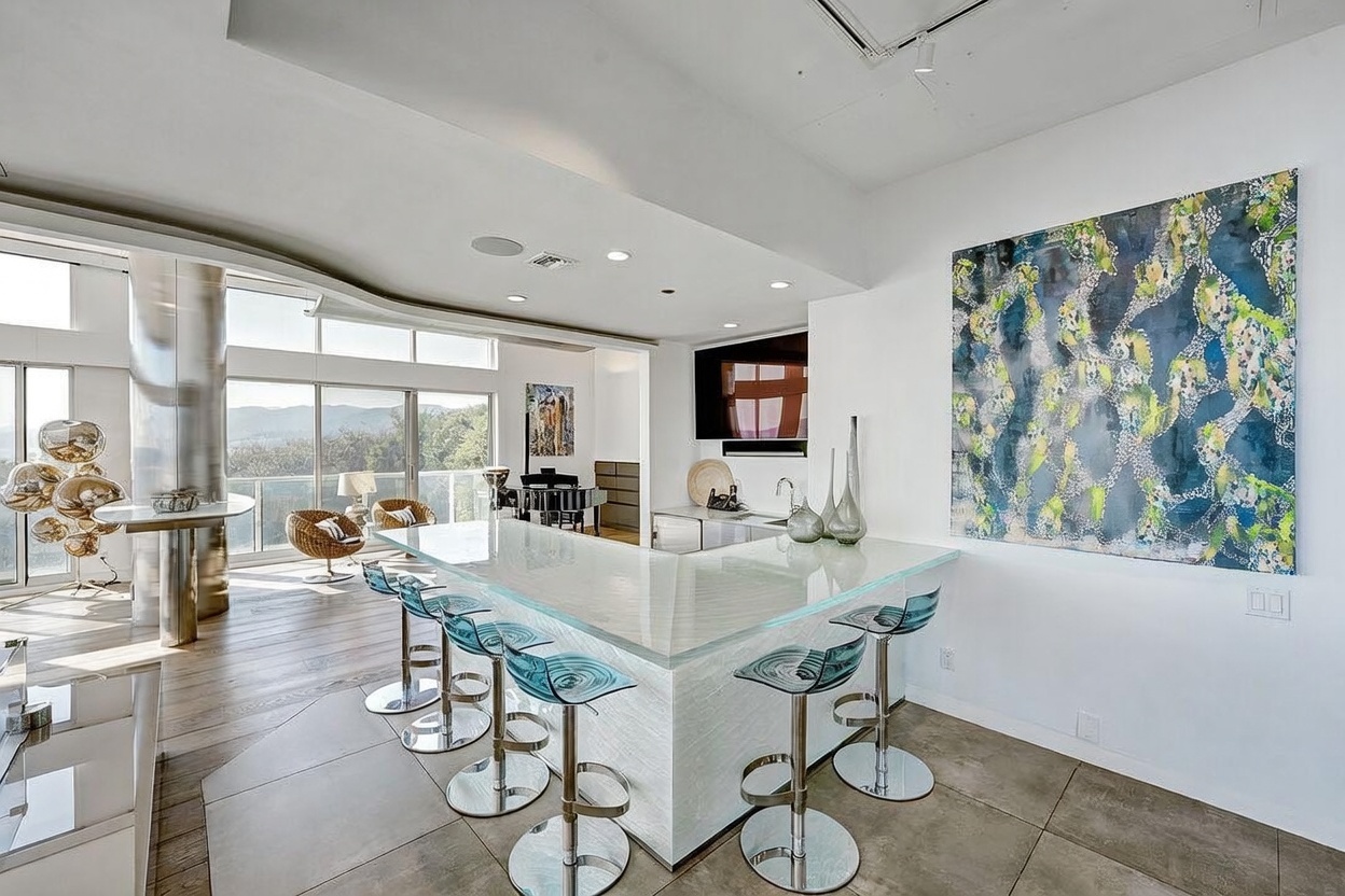 This modern, open-concept kitchen features a large, L-shaped island with a translucent glass countertop and a textured white base, surrounded by sleek, blue-tinted acrylic bar stools. The space is bright and airy, with light wood flooring, a large abstract painting on the wall, and expansive windows that provide a panoramic view of the surrounding hills. A secondary seating area with wicker chairs and a piano is visible in the background, emphasizing the home's spacious and sophisticated layout.