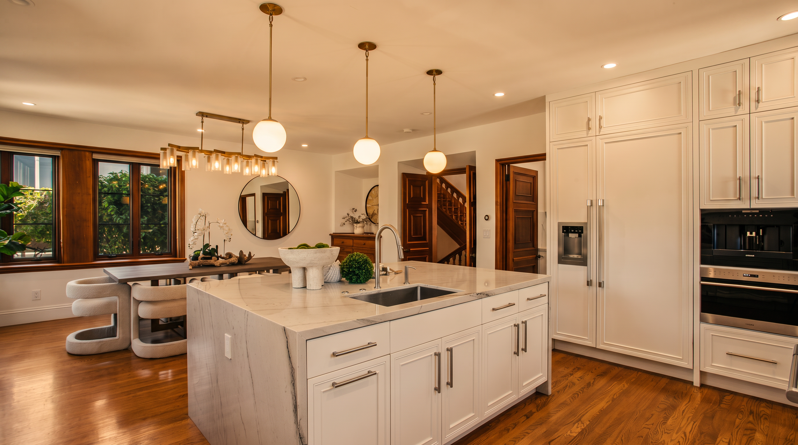 This bright and spacious kitchen features a large central island with a stone countertop and an integrated sink, illuminated by three modern pendant lights. The space is anchored by white cabinetry, high-end built-in appliances, and warm wood flooring that extends into the adjacent dining area. The open-concept layout creates a seamless flow, highlighted by a large dining table and a view into the hallway with a staircase.