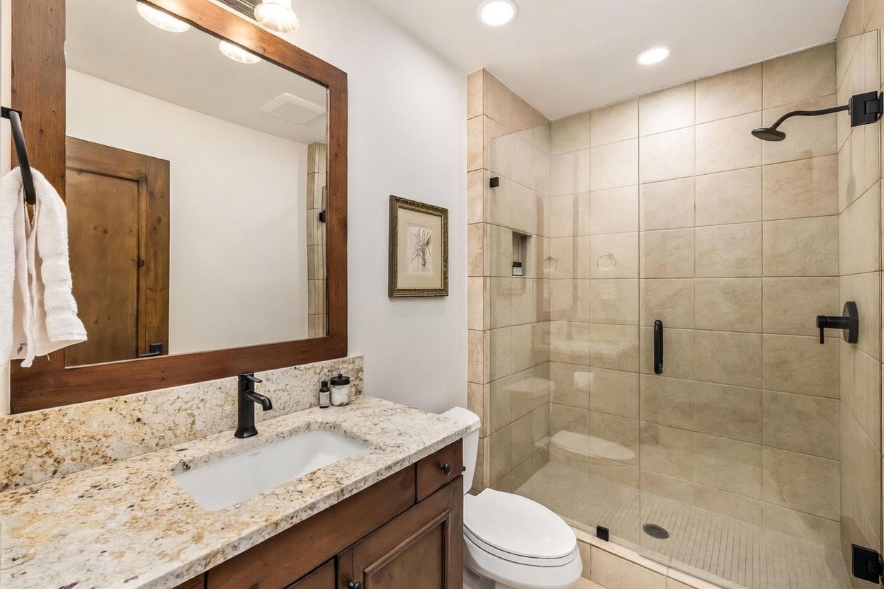 This bathroom features a vanity with a stone countertop, a dark wood cabinet, and a framed mirror above the sink. To the right, a glass-enclosed shower is finished with neutral-toned wall tiles and includes a built-in recessed shelf. The space is completed by a white toilet and modern dark-toned fixtures, creating a cohesive and functional design.